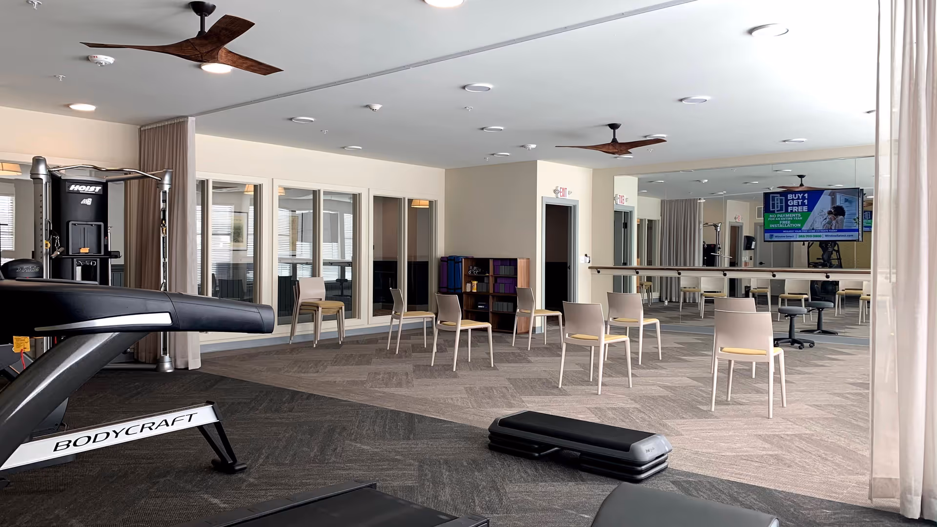 Spacious interior fitness/activity room with a treadmill, step platforms, chairs arranged in a circle, mirrored wall and a wall-mounted TV.