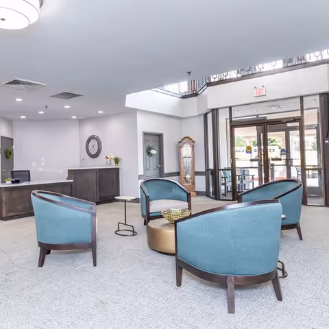 A bright and spacious lobby area with four teal upholstered chairs arranged around a round gold coffee table. The room features a high ceiling with a large window above the entrance doors, a reception desk to the left, a wall clock, and a grandfather clock near the entrance. The floor is carpeted in a light color, and the entrance doors have glass panels allowing natural light to enter.