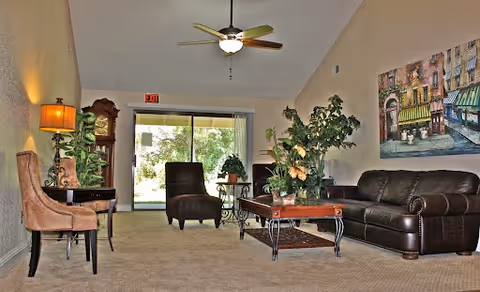 Spacious communal living room with a leather sofa, chairs, coffee table, plants, ceiling fan, and sliding glass doors to the outside.