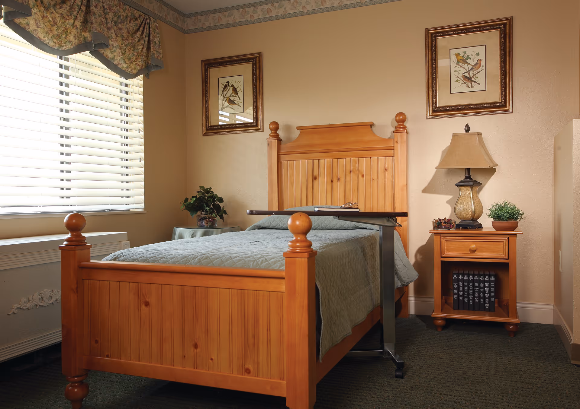 A cozy bedroom with a wooden bed frame and a green quilted bedspread. There is a small overbed table on wheels positioned over the bed. Next to the bed is a wooden nightstand with a lamp, a small potted plant, and some decorative items. Two framed bird illustrations hang on the beige walls above the bed. A window with white blinds and a floral valance lets in natural light.