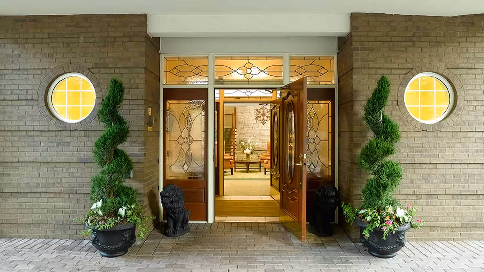 Entrance to a building with double wooden doors featuring decorative glass panels. Two spiral-shaped topiary plants in large black pots flank the doorway, each accompanied by a black lion statue. The building exterior is made of light brown brick with two round windows on either side of the entrance. Inside, a seating area with chairs and a coffee table with a flower arrangement is visible.