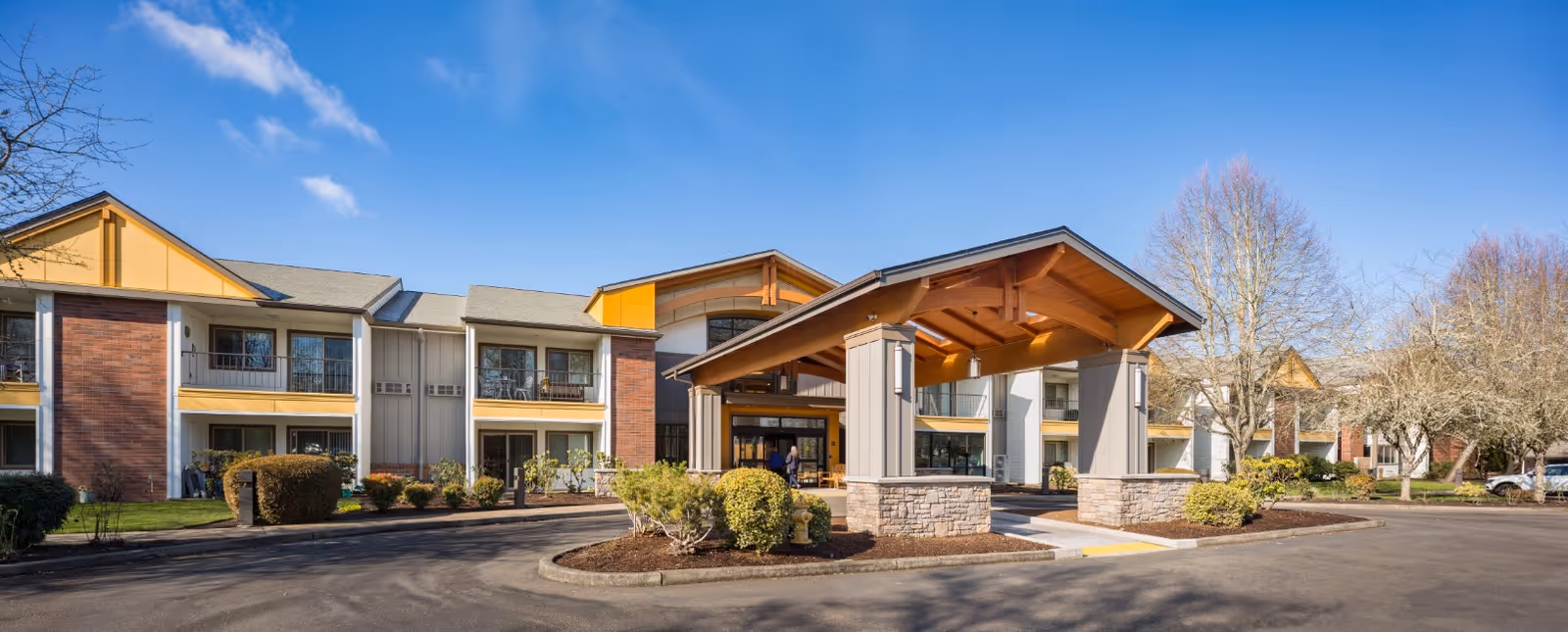 Exterior view of Jennings McCall Center, a two-story senior living facility with a covered entrance, landscaped bushes, and leafless trees under a clear blue sky.