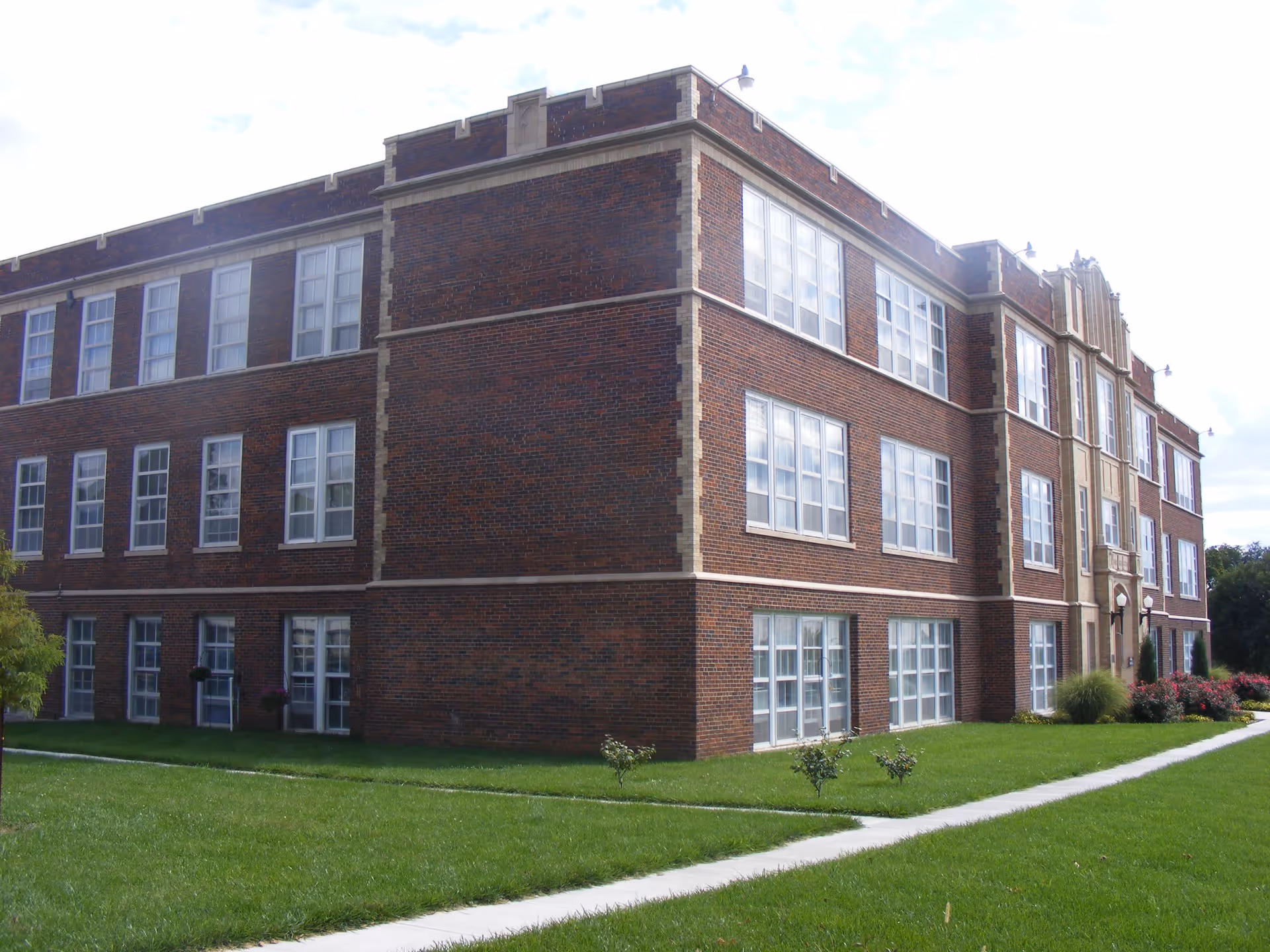 Exterior view of a three-story brick building with many large windows, surrounded by a well-maintained green lawn and a concrete walkway. The building has decorative stone accents and some landscaping with small bushes and flowers near the entrance.