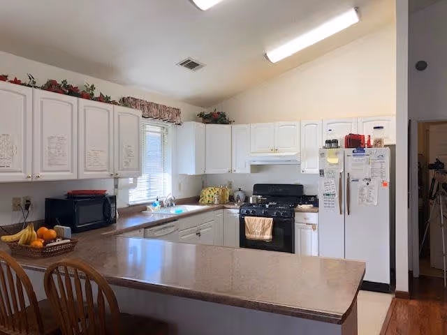A bright kitchen with white cabinets, a beige countertop, a black microwave, a white refrigerator covered with notes and magnets, a black stove with a beige towel hanging on the handle, and a window above the sink letting in natural light. There are two wooden chairs at the counter and a basket of fruit on the counter.