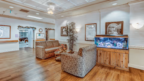 A cozy seating area in a senior living facility with two patterned armchairs and a small round table with flowers. There is a large fish tank on a wooden stand to the right, framed artwork and a tall plant on the wall behind the chairs. The room has wood flooring, white walls with wainscoting, and a ceiling fan with lights. An open doorway leads to another room with tables and chairs.