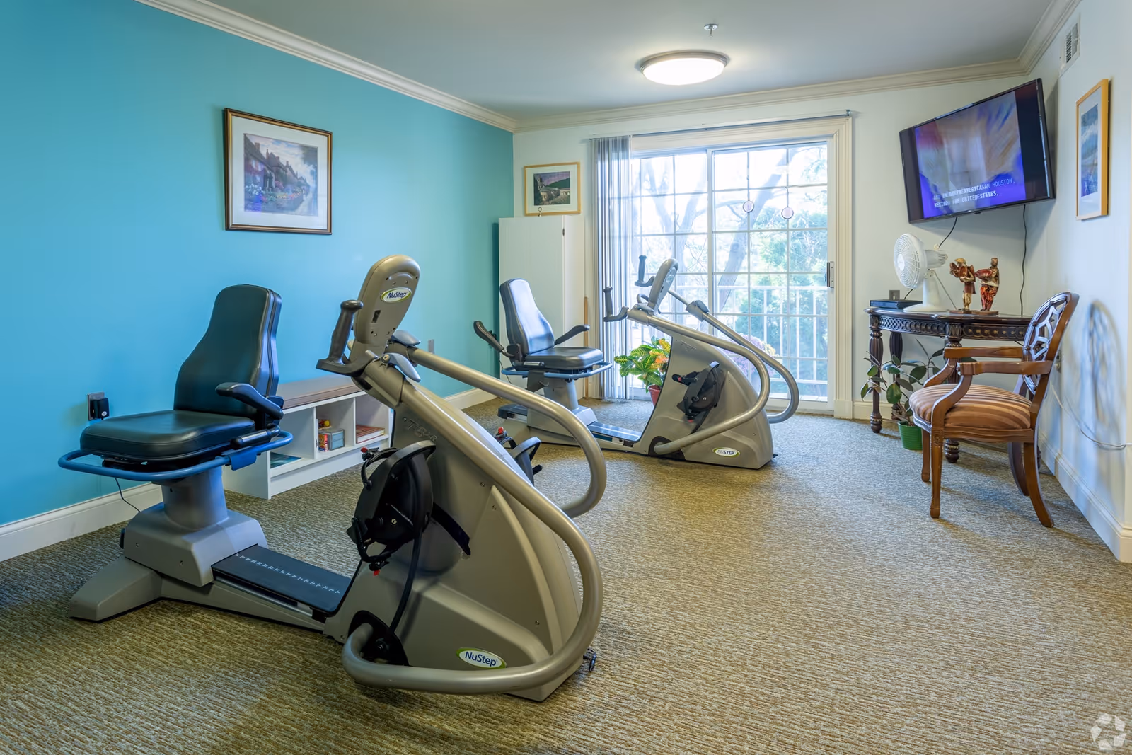 A small exercise room with two NuStep recumbent cross trainers, a wooden chair, a small table with decorative items, a wall-mounted TV, and a large window with vertical blinds letting in natural light. The walls are painted blue and white, and there are framed pictures hanging on the walls.