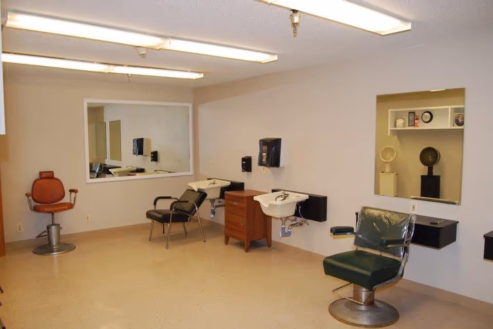 Interior of a hair salon area with three salon chairs, two white sinks mounted on the wall, large mirrors, and shelves with hair drying equipment. The room has beige walls and a tiled floor with fluorescent ceiling lights.