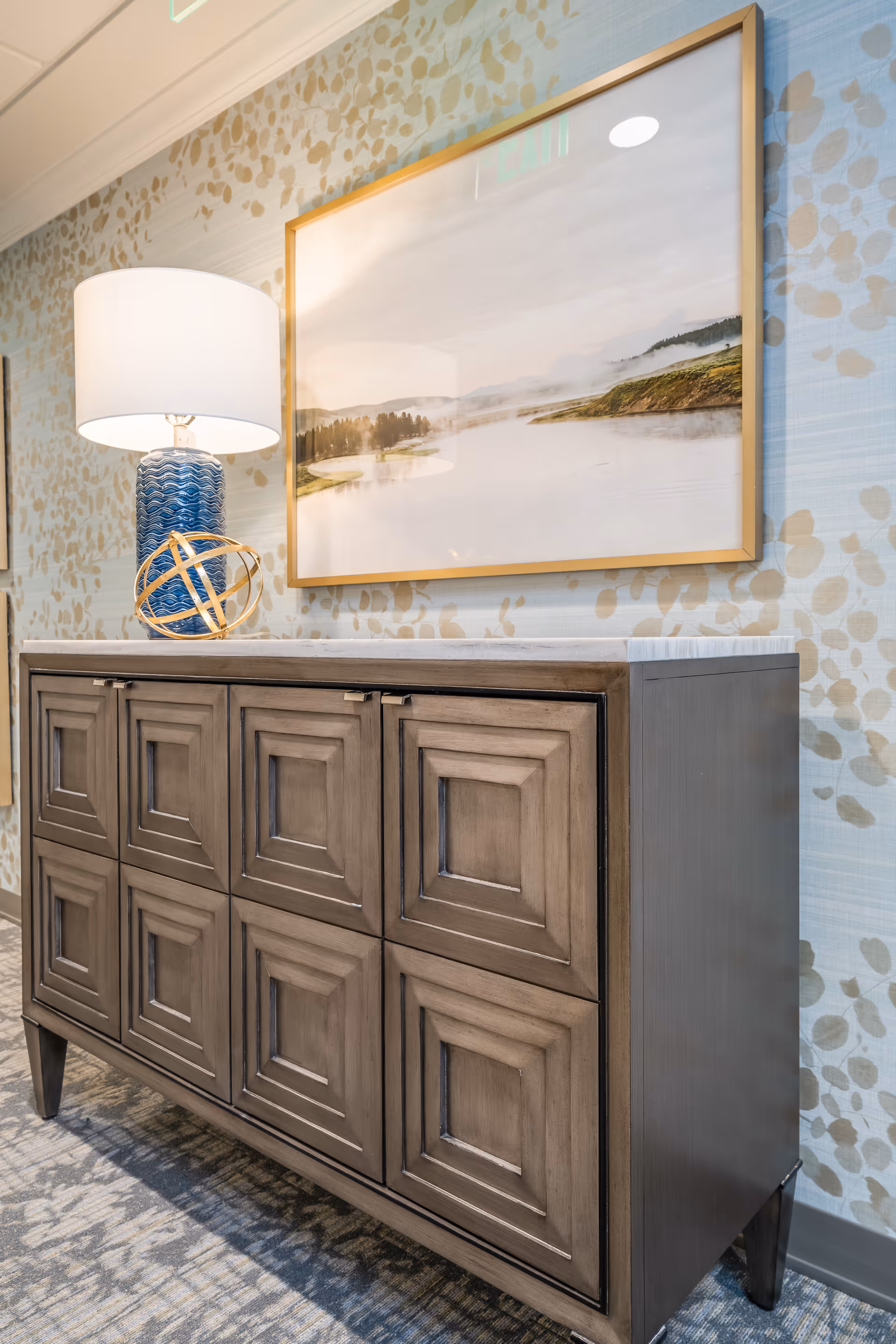 A wooden cabinet with decorative paneling stands against a wall with patterned wallpaper. On top of the cabinet is a blue table lamp with a white shade and a gold decorative orb. Above the cabinet hangs a framed landscape painting depicting a misty lake scene with trees and hills.
