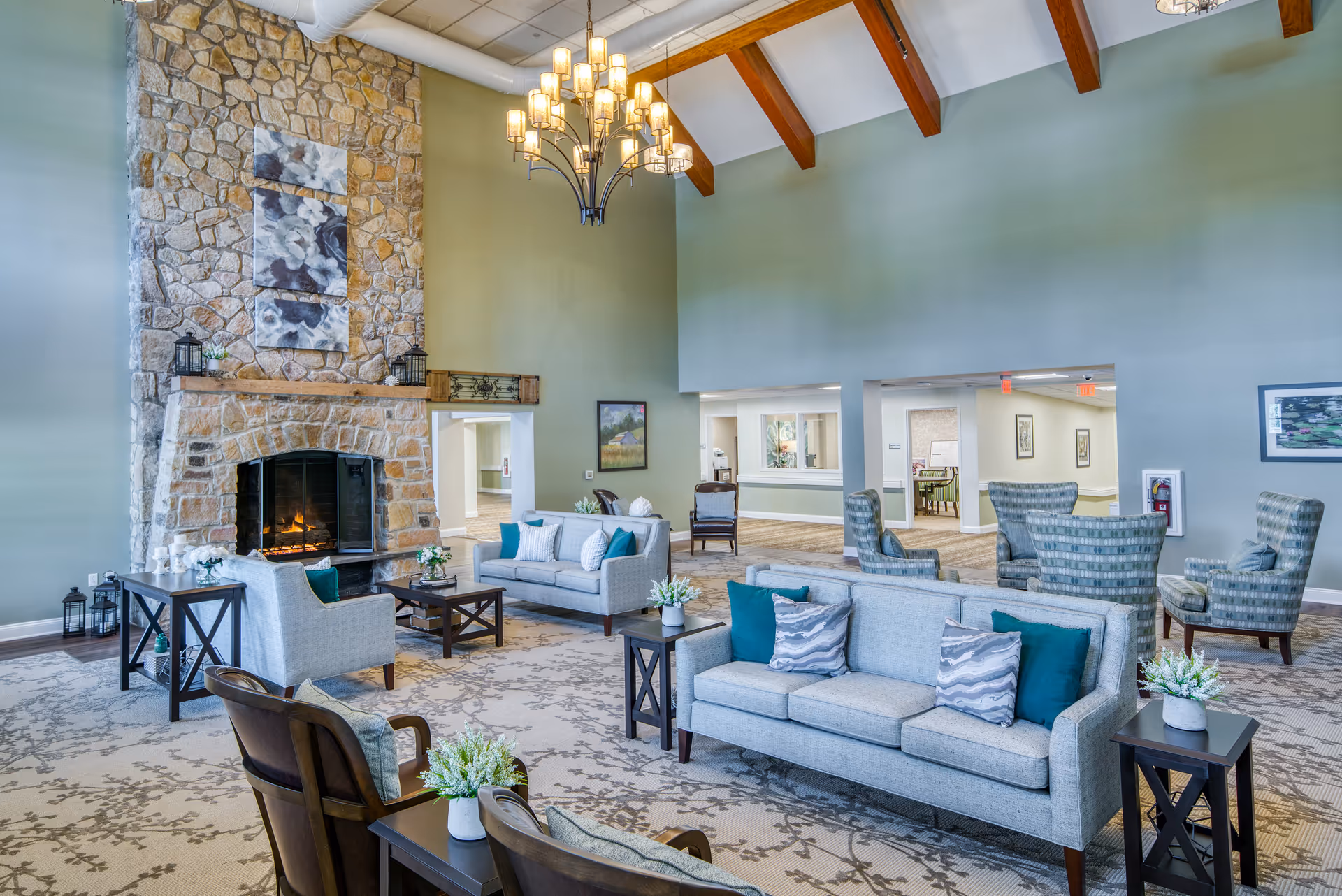 Spacious senior living facility common area with high vaulted ceiling and wooden beams, featuring a large stone fireplace with a fire burning, multiple seating arrangements including sofas and armchairs with cushions, side tables with flower vases, and a chandelier hanging from the ceiling.