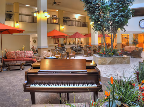 Indoor atrium of a senior living facility with a grand piano in the foreground, seating areas with umbrellas, plants and upper-level balconies.