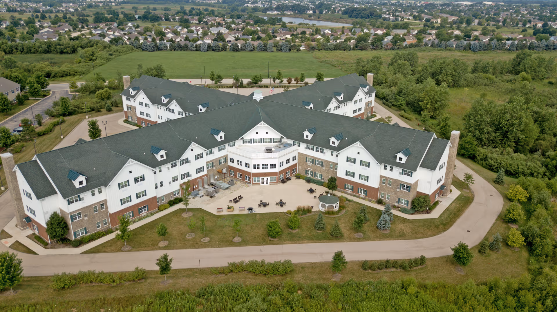 Aerial view of a large senior living facility building with multiple wings, surrounded by greenery and trees. The building has a green roof and white exterior walls with brick accents. There is a paved driveway and outdoor seating areas visible in the courtyard. Residential neighborhoods and open fields are seen in the background.