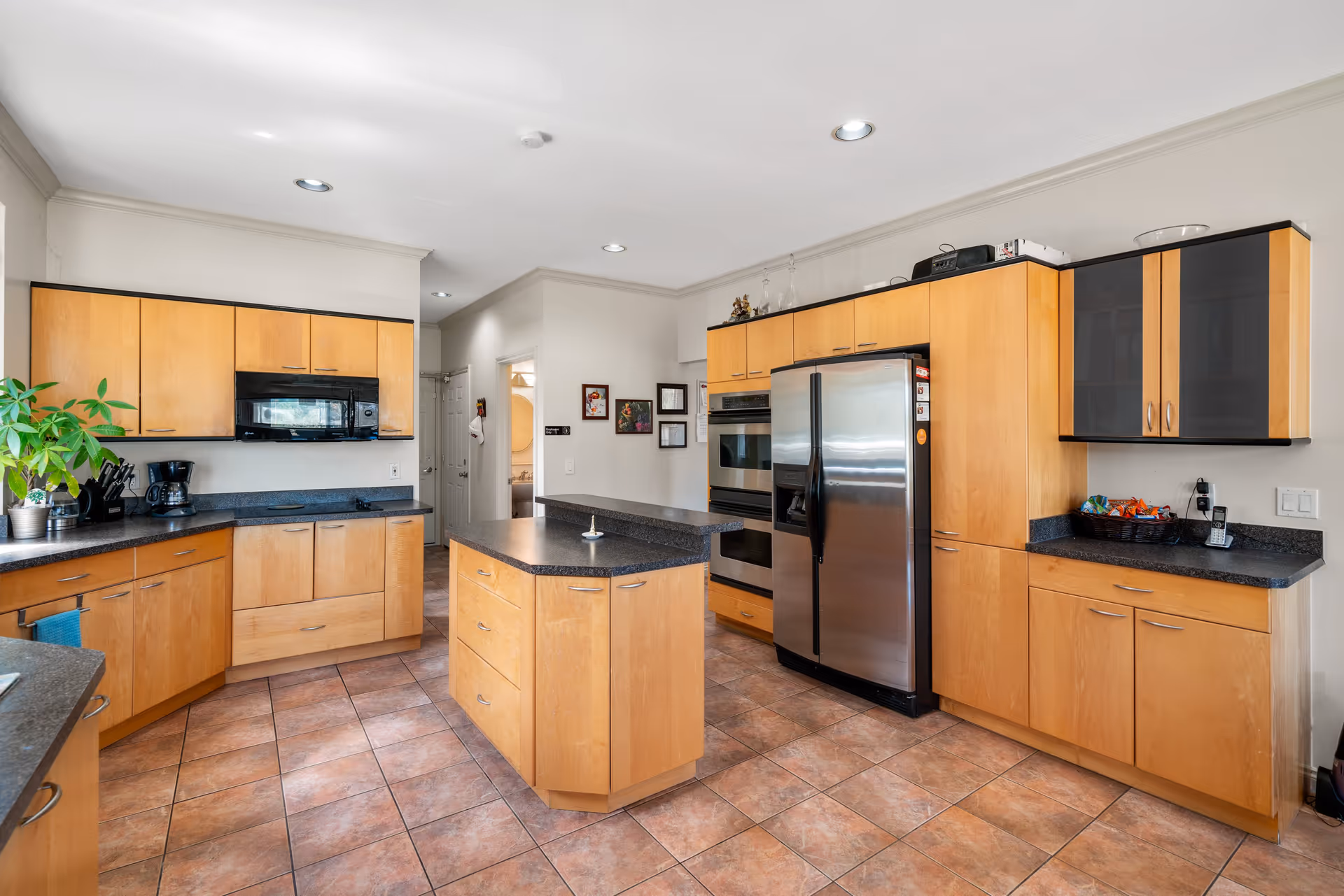 A spacious kitchen with light wood cabinets, black countertops, and tiled floor. The kitchen features a central island with drawers, a stainless steel refrigerator, built-in double ovens, a black microwave above the stove, and various small appliances and decorations on the counters. The walls are painted white, and there are recessed ceiling lights.