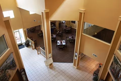 View from an upper level overlooking a spacious interior area with tiled flooring and beige walls. The area below features multiple tables and chairs arranged in a dining setup, with large windows allowing natural light to enter.