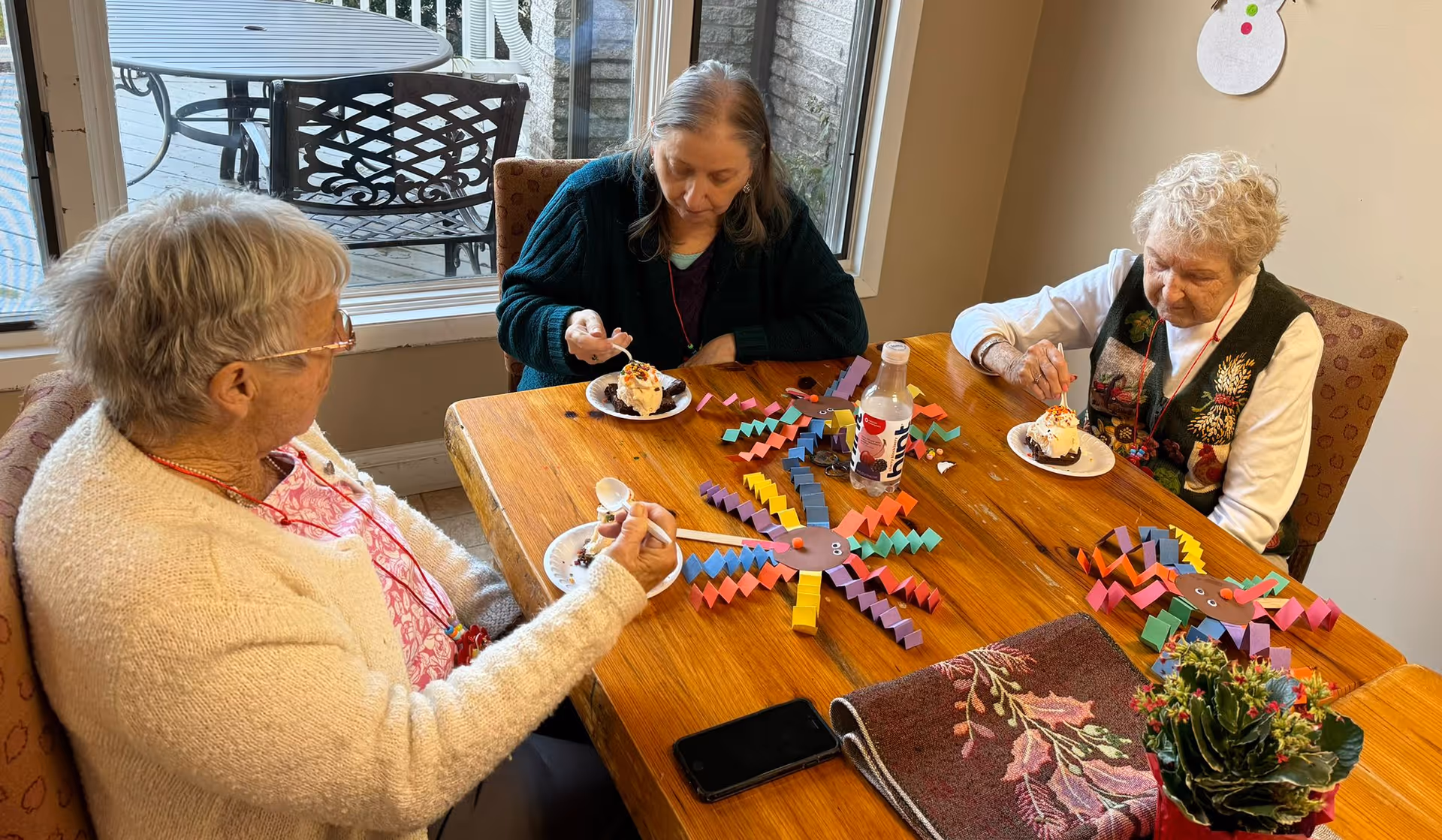 Three elderly women sitting around a wooden table inside a room, eating dessert from small plates. The table has colorful paper crafts shaped like spiders and a small plant. There is a window behind them showing outdoor patio furniture.