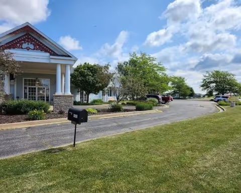 Exterior view of a senior living facility with a covered entrance supported by white columns, a mailbox on a post near the driveway, several parked cars, green grass, trees, and a partly cloudy sky.