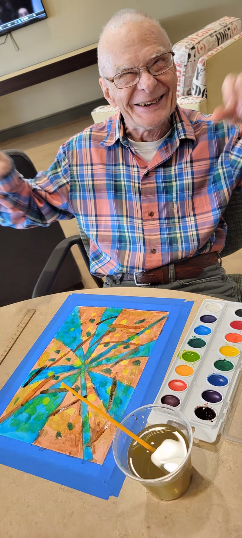 An elderly man wearing glasses and a colorful plaid shirt is smiling and raising his arms while sitting at a table. On the table in front of him is a watercolor painting with abstract shapes in blue, green, and orange, a paintbrush resting in a cup of water, and a watercolor paint palette.
