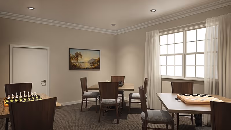 A well-lit interior room with beige walls and carpeted floor featuring three wooden tables each surrounded by wooden chairs with cushioned seats. Two tables have board games set up, one with a chessboard and the other with a checkers board. A large window with sheer white curtains allows natural light into the room. A landscape painting hangs on the wall near a closed white door.