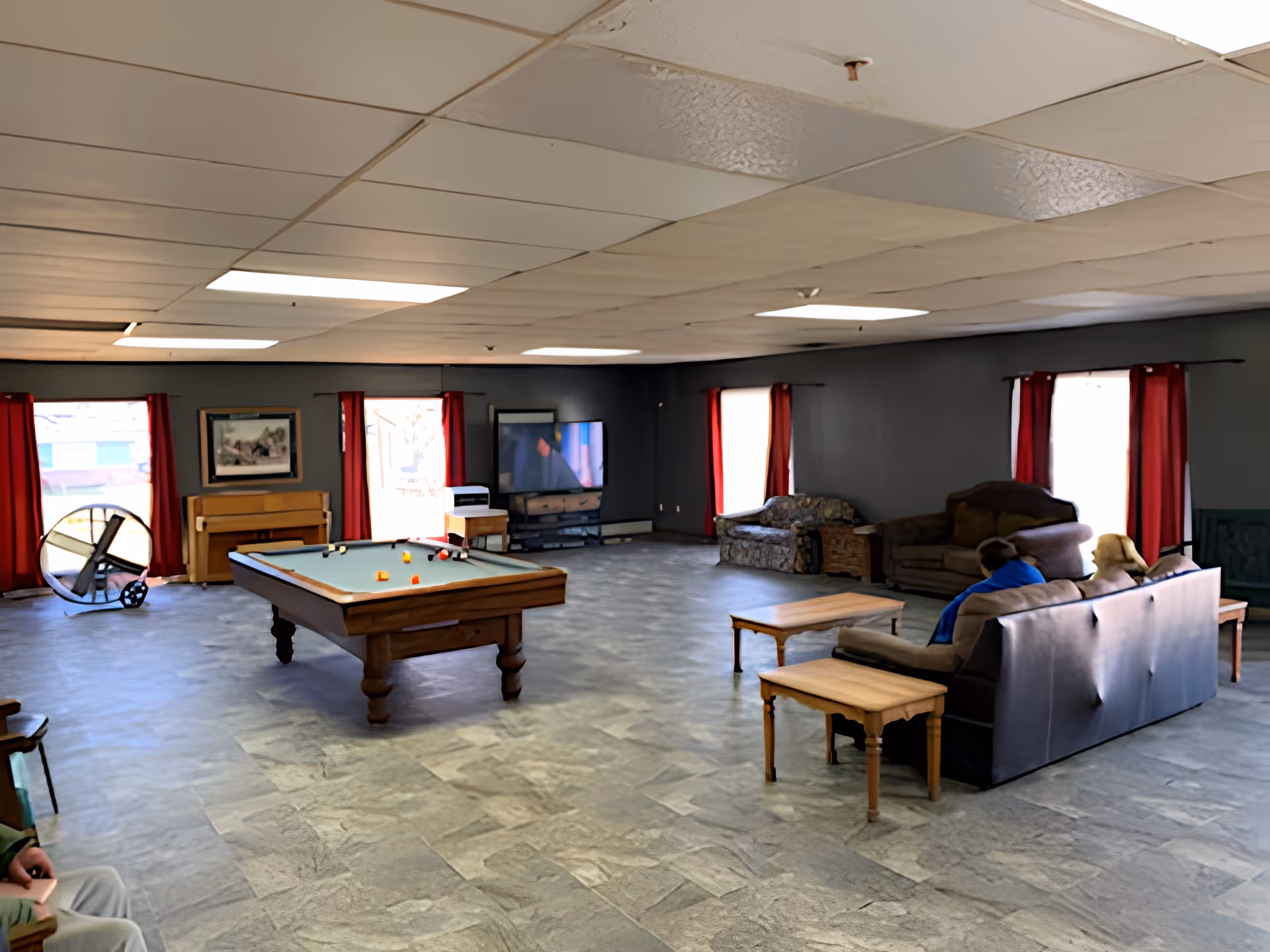 Spacious communal lounge with a pool table in the center, couches and a TV near windows with red curtains.