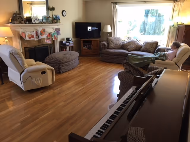 A cozy living room with wooden flooring featuring a beige recliner chair, a patterned sofa with cushions, a matching ottoman, a piano in the foreground, a fireplace with decorative items on the mantel, a TV on a wooden stand, and a large window with curtains letting in natural light. A person is sitting on the recliner covered with a green blanket.