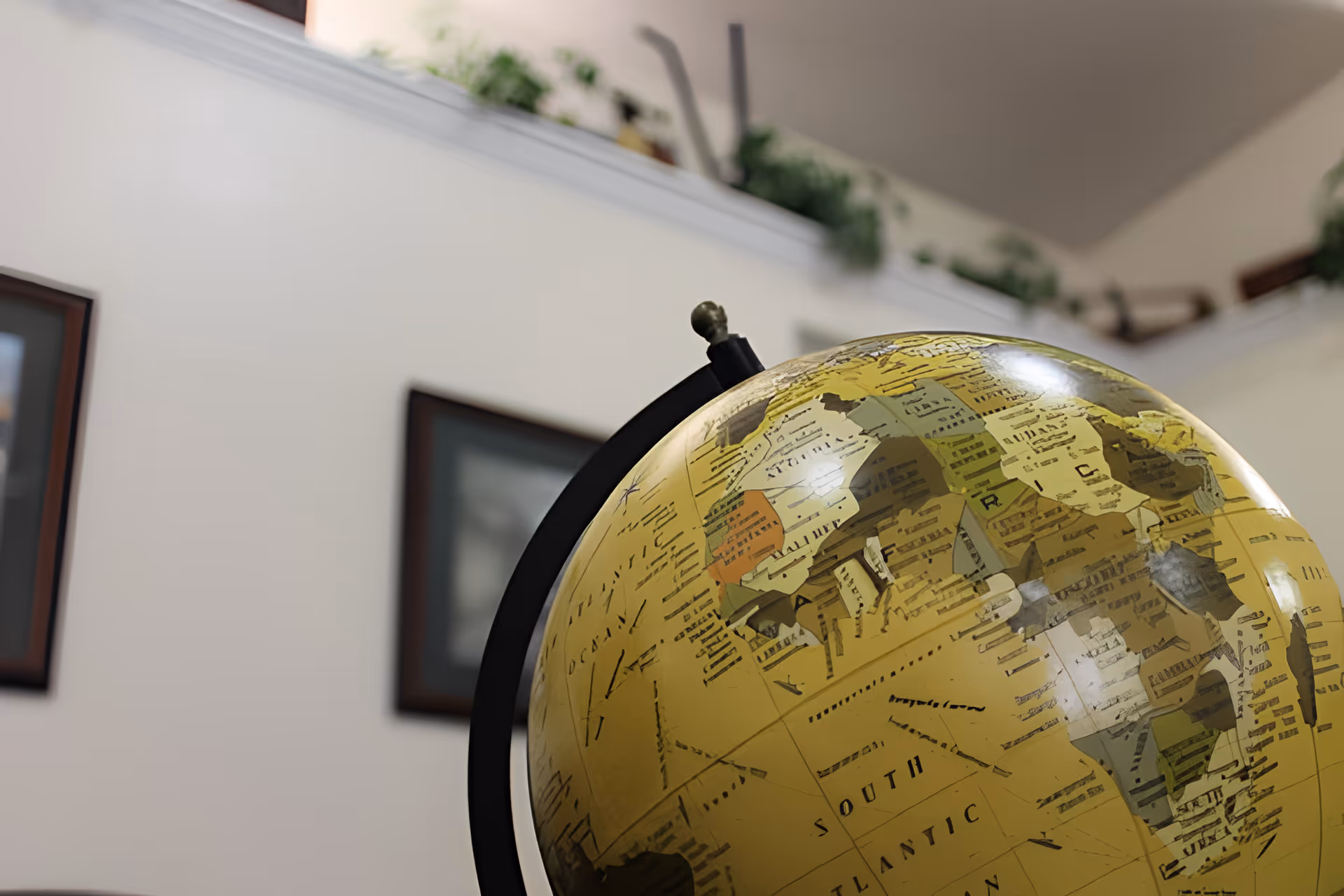 Close-up view of a yellow globe showing parts of Africa and the South Atlantic Ocean, with a blurred background of a white wall, framed pictures, and green plants on a high shelf.
