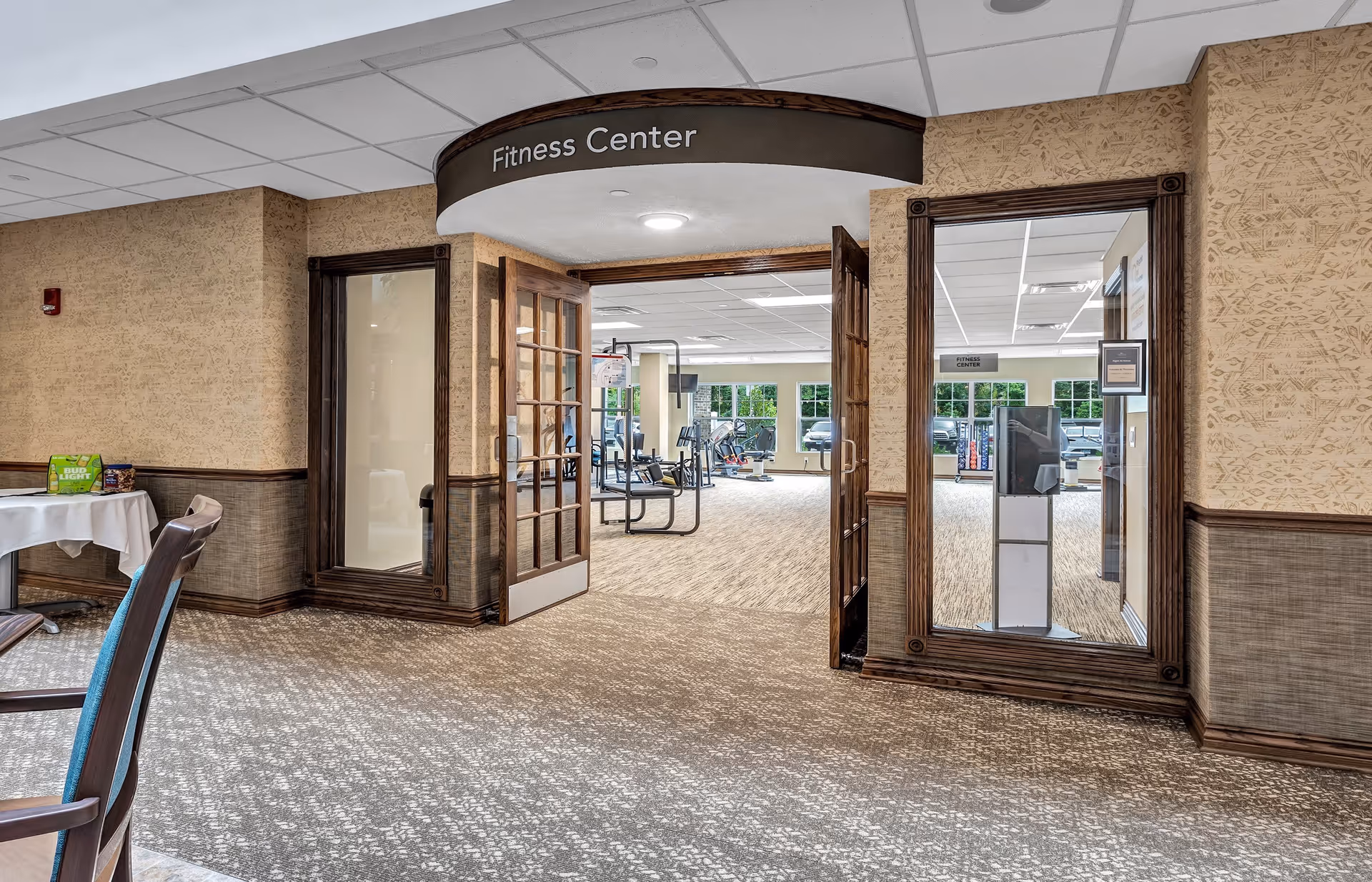 Entrance to a fitness center in a retirement community with double wooden doors and glass panels. Inside, exercise equipment is visible. The area outside the fitness center has carpeted flooring and a table with chairs.