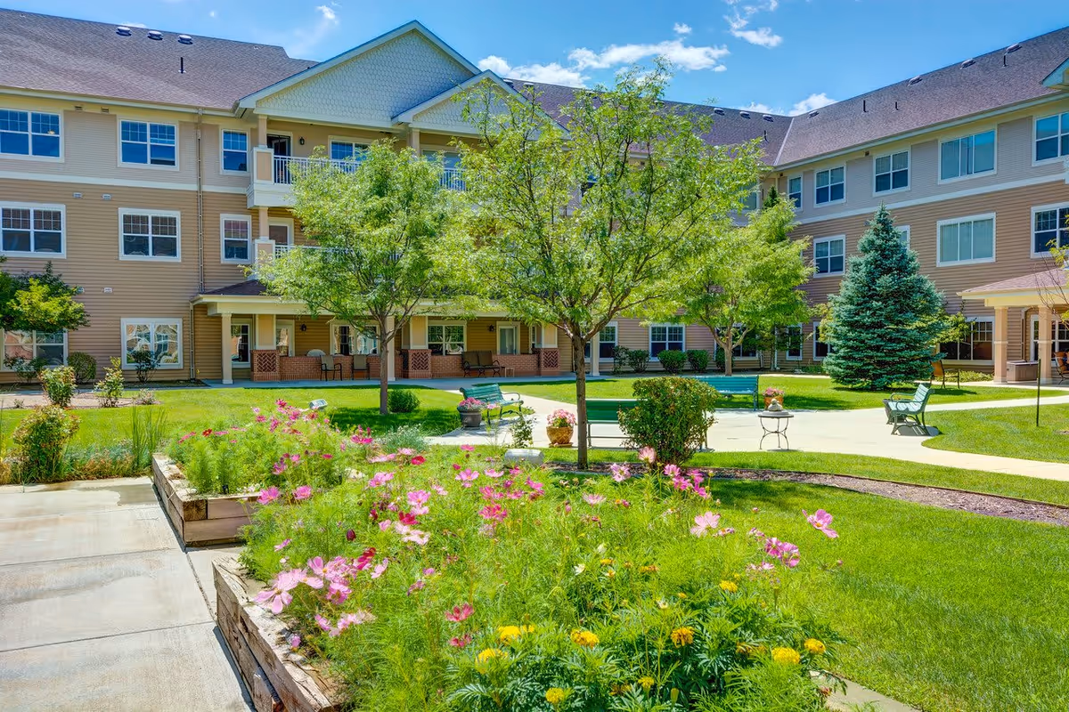 A bright and sunny outdoor courtyard at The Courtyards at Mountain View featuring green grass, blooming pink and yellow flowers in raised garden beds, several trees, benches, and a three-story beige building with multiple windows in the background.