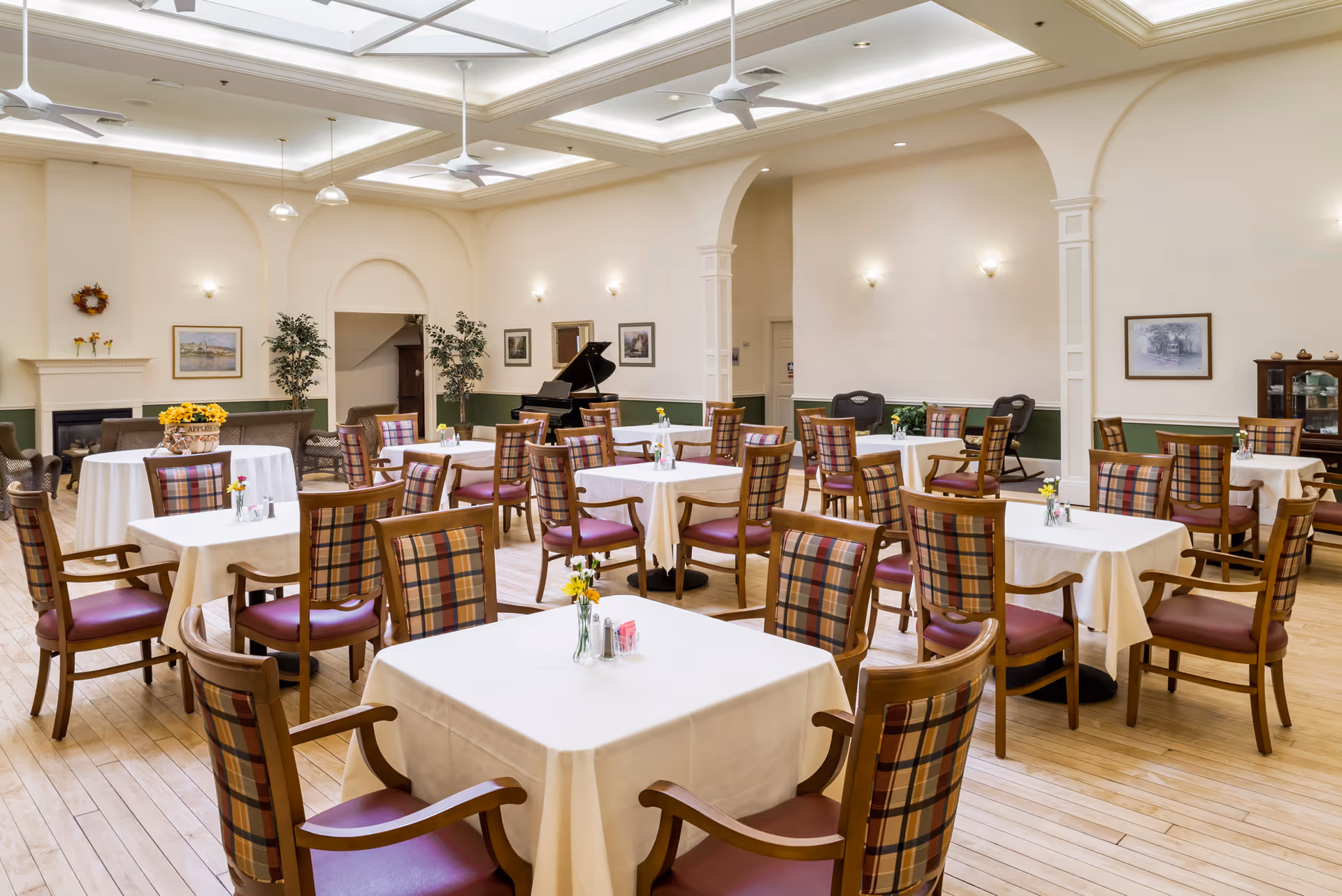 Spacious, well-lit dining room with multiple tables draped in white cloths, plaid-backed wooden chairs, a grand piano and fireplace in the background.