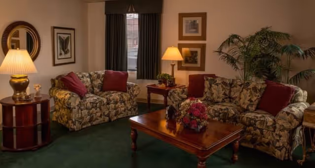 A cozy living room with two floral-patterned sofas adorned with red throw pillows, a wooden coffee table with a flower arrangement, a side table with a lamp and decorative items, framed pictures on the wall, a round mirror, and a large potted plant near the corner window with dark curtains.