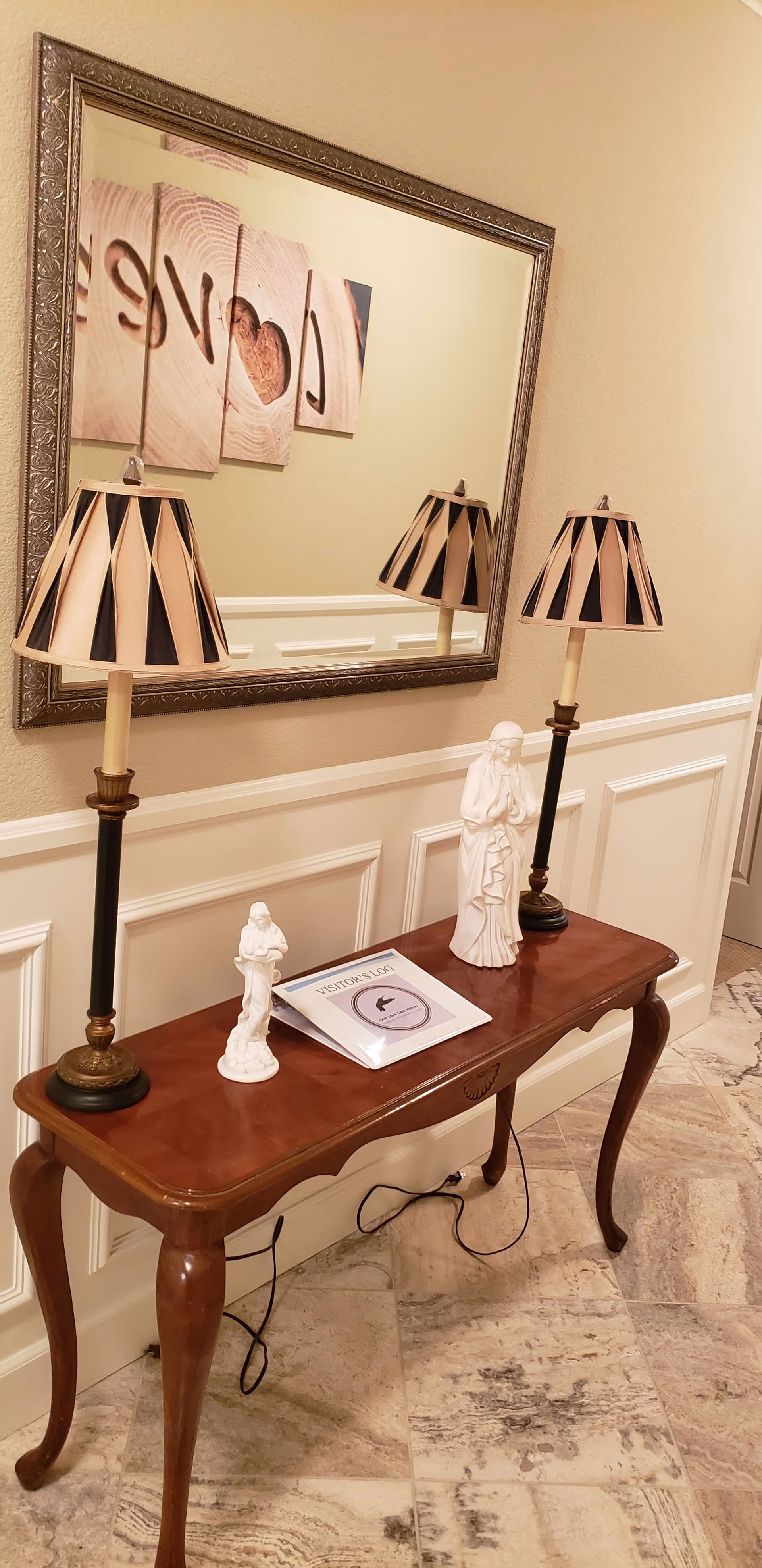A wooden console table with curved legs placed against a beige wall with white wainscoting. On the table are two decorative lamps with black and beige striped shades, two white statues, and a booklet titled 'VISITORS LIST'. Above the table is a large rectangular mirror with an ornate frame reflecting part of a wall art piece that spells 'Love' with a heart shape.