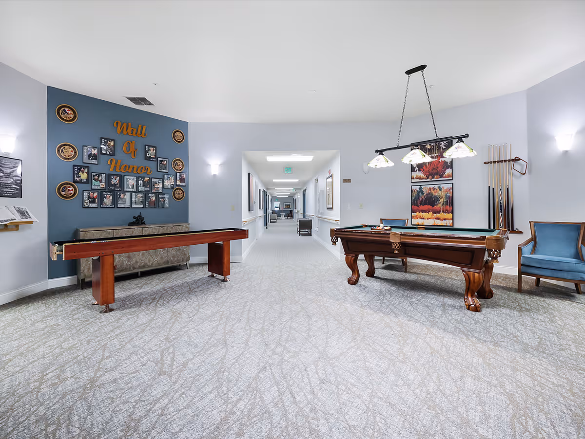 A recreational area in a senior living facility featuring a shuffleboard table on the left and a pool table with hanging lights on the right. The back wall has a 'Wall of Honor' display with photos and plaques. The space has light-colored walls and carpeted flooring, with a hallway extending in the background.
