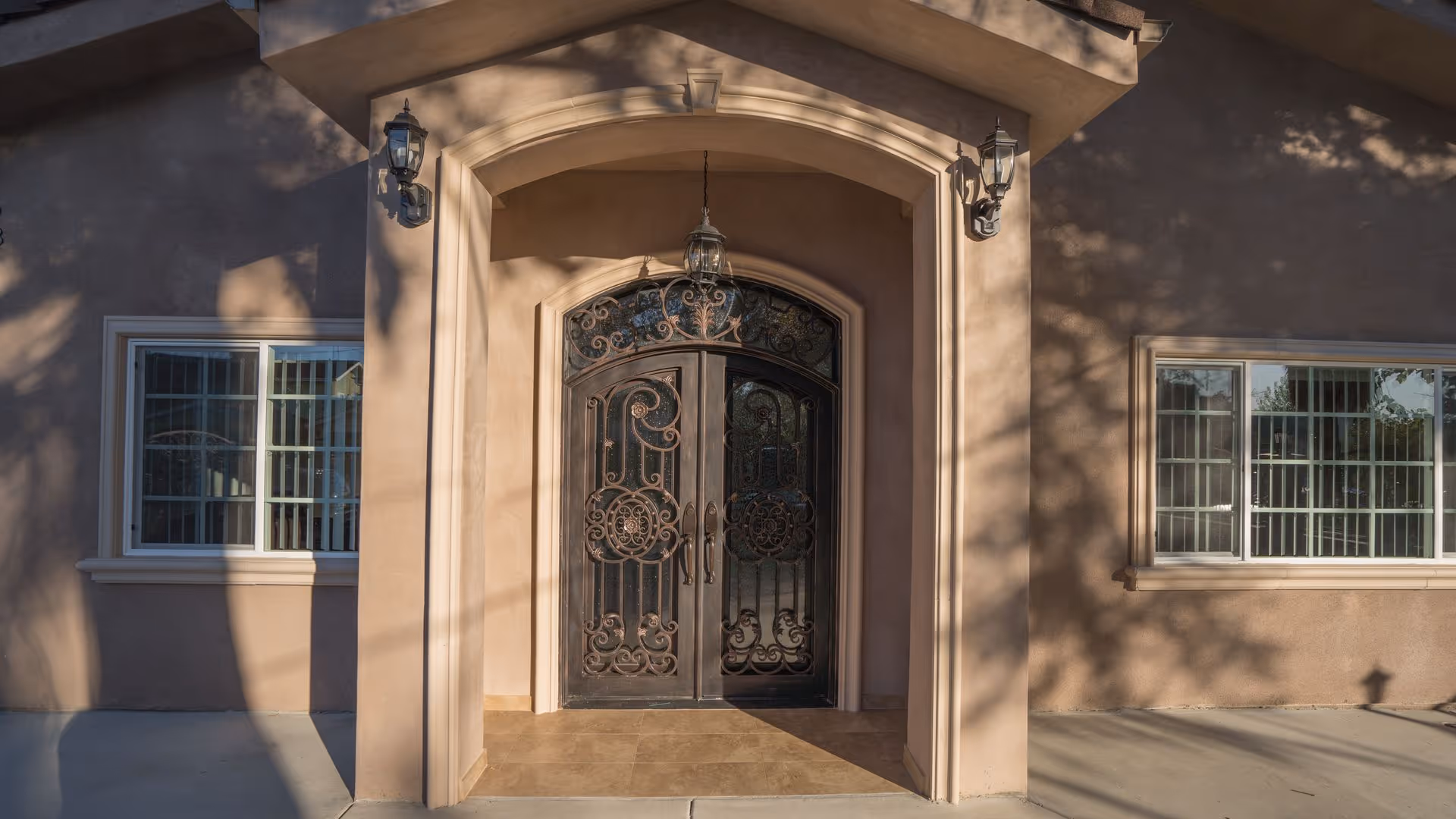 Arched front entrance of a building with ornate wrought-iron double doors, flanked by windows and exterior wall lanterns.