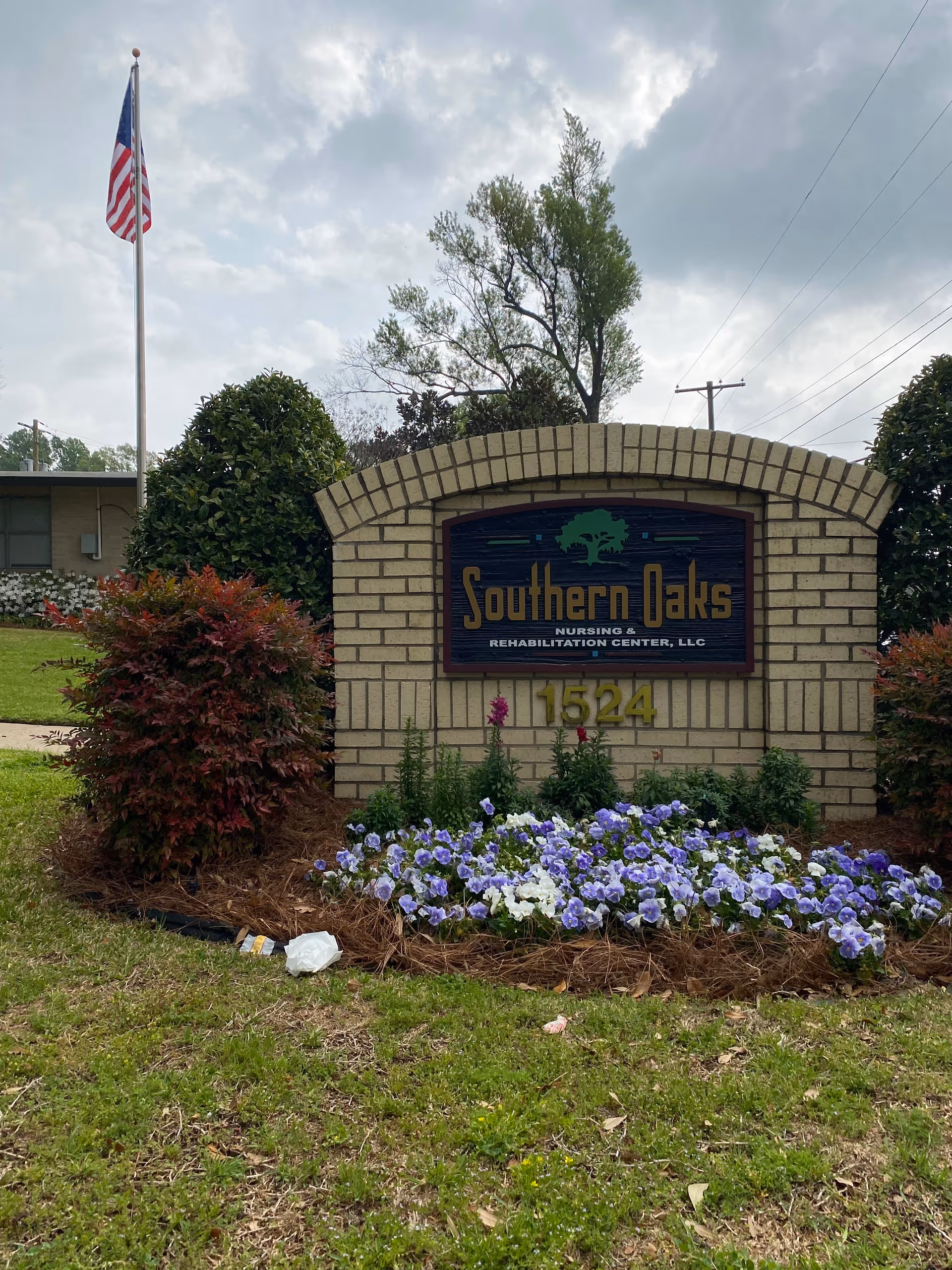 A brick sign for Southern Oaks Nursing & Rehabilitation Center, LLC with the address number 1524. The sign is surrounded by purple and white flowers, bushes, and grass. An American flag on a flagpole is visible in the background under a cloudy sky.