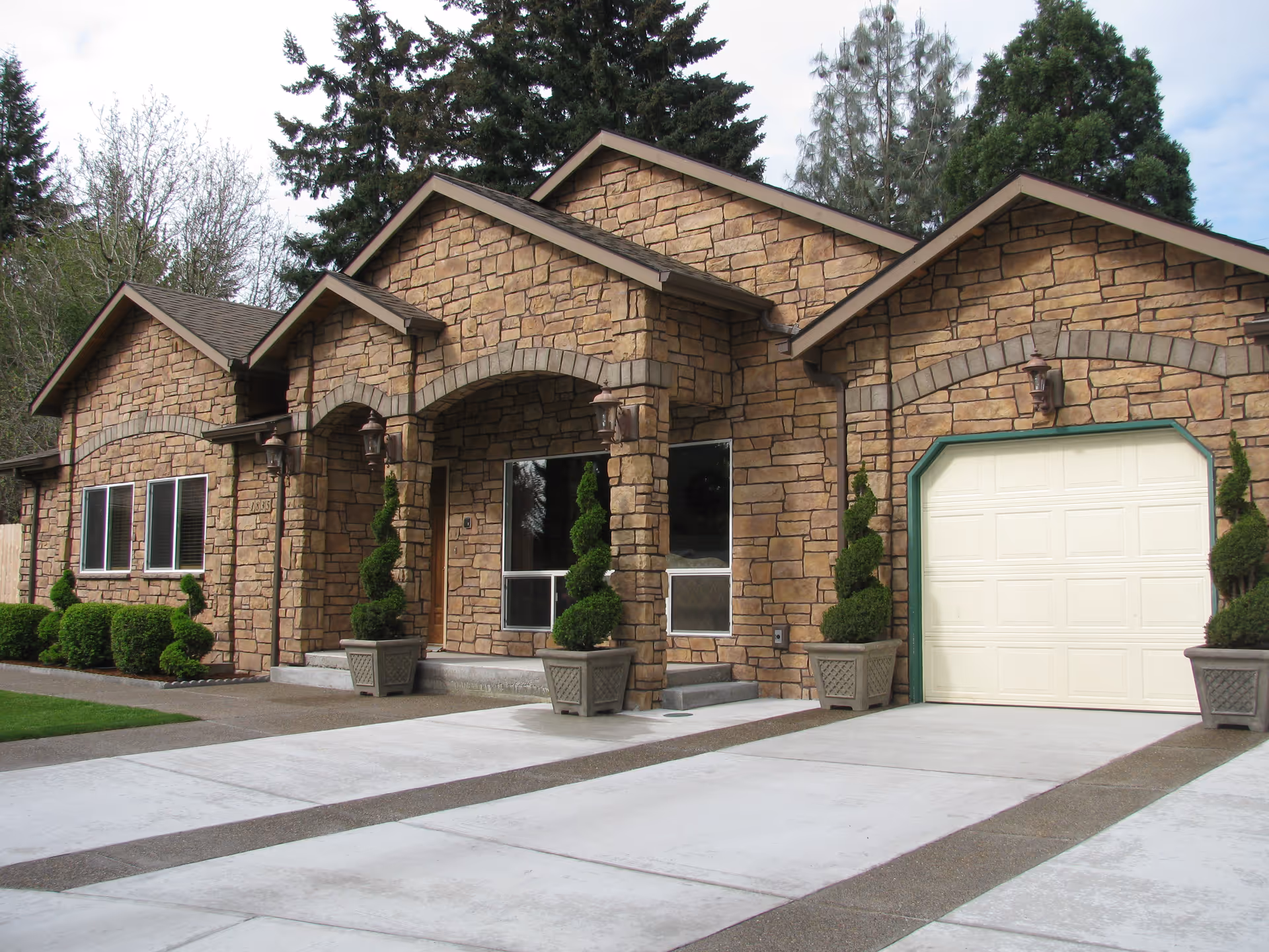 Exterior view of a stone-faced building with a garage door, multiple windows, and a covered entrance with decorative topiary plants in large planters. The driveway and walkway are concrete, and there are trees in the background.