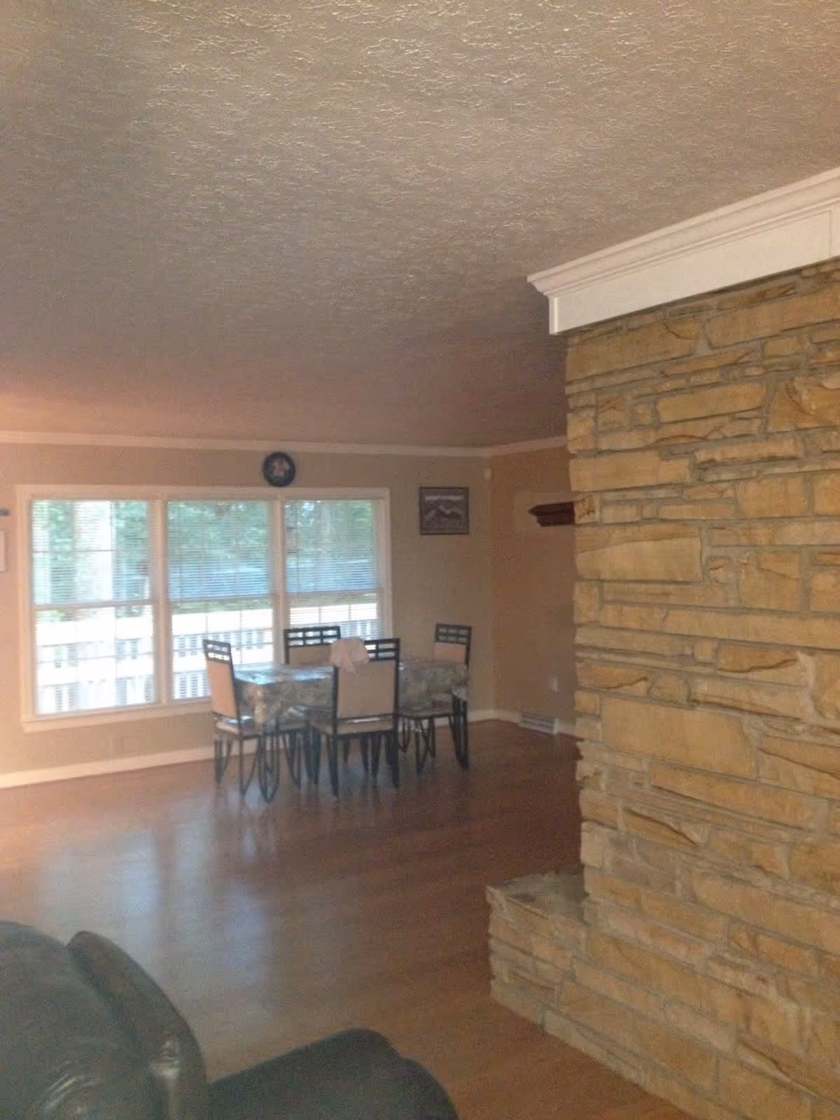 Interior view of a living area with a stone fireplace on the right, wooden flooring, a dining table with six chairs near a large window with blinds, and beige walls.