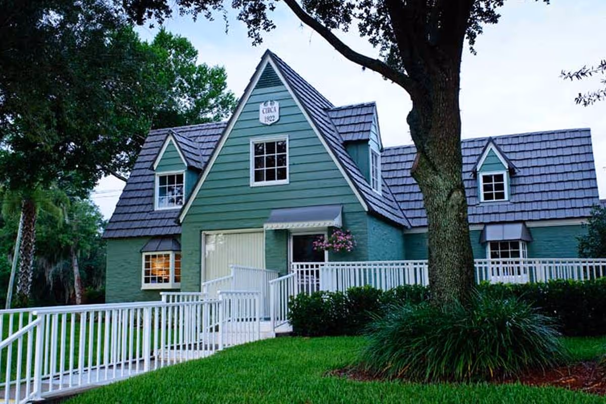 Exterior view of a green building with a steeply pitched roof and multiple dormer windows. There is a white railing ramp leading to the entrance, surrounded by green grass, bushes, and trees. A sign on the building reads 'Circa 1922'.