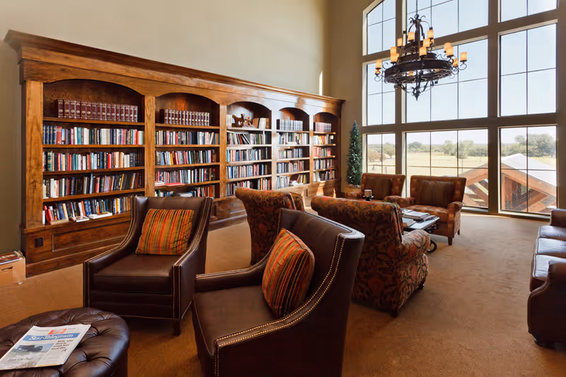 A spacious and well-lit library or lounge area with large wooden bookshelves filled with books along one wall. There are several comfortable armchairs with patterned cushions arranged around a coffee table. A large window with multiple panes lets in natural light and offers a view of the outdoors. A decorative chandelier hangs from the ceiling.