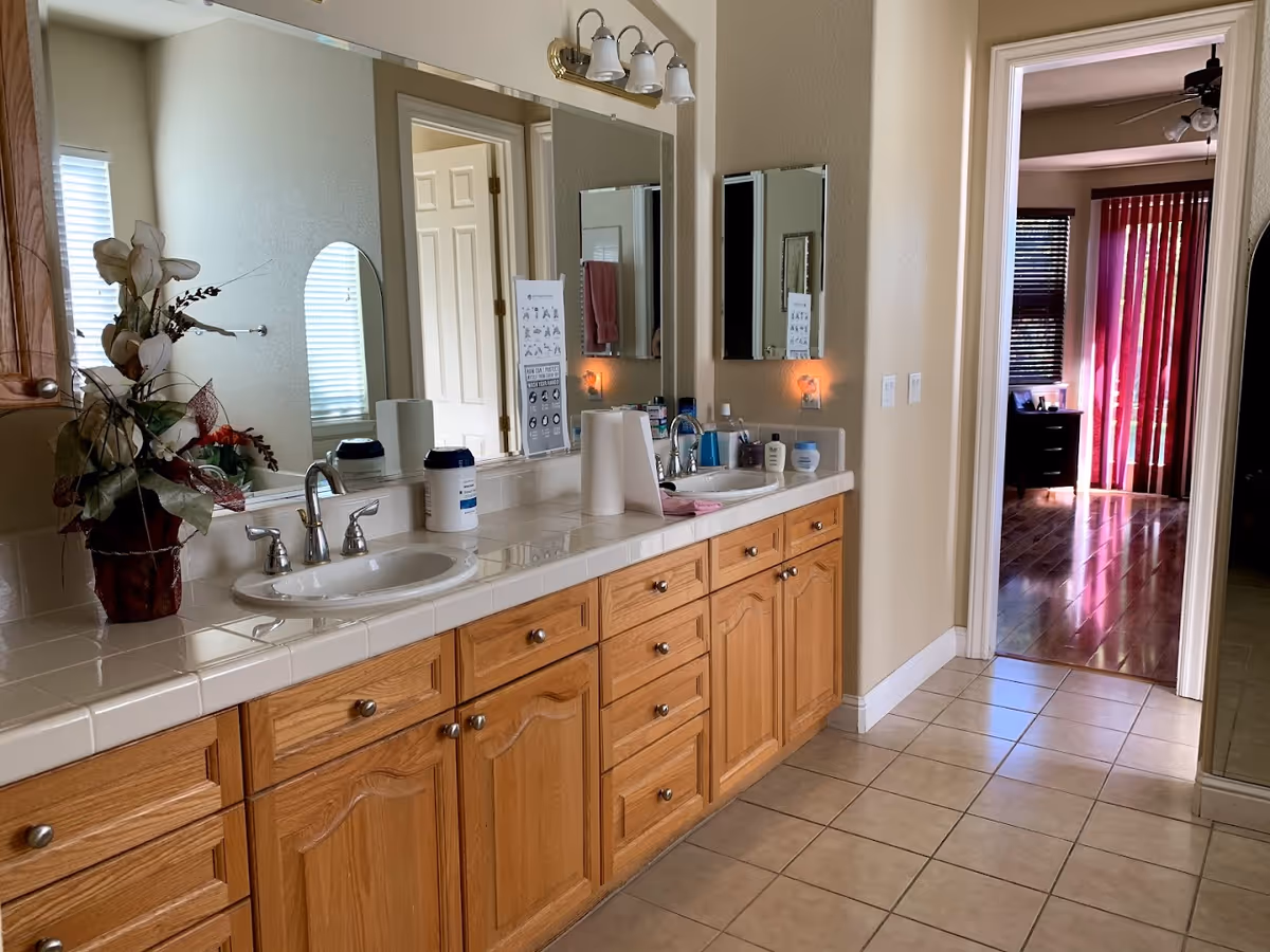 A bathroom with a long countertop featuring two sinks and wooden cabinets underneath. The countertop has various items including a flower arrangement, paper towels, and personal care products. Above the sinks is a large mirror with a light fixture. The bathroom floor is tiled, and there is a doorway leading to a room with wooden flooring and red vertical blinds.