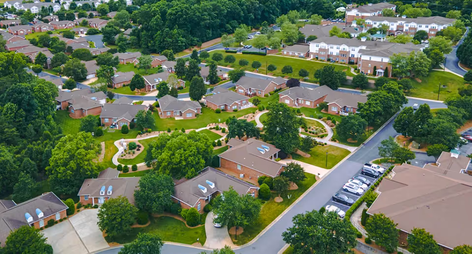 Aerial view of a senior living campus with brick cottages, winding walkways, trees, and parked cars.