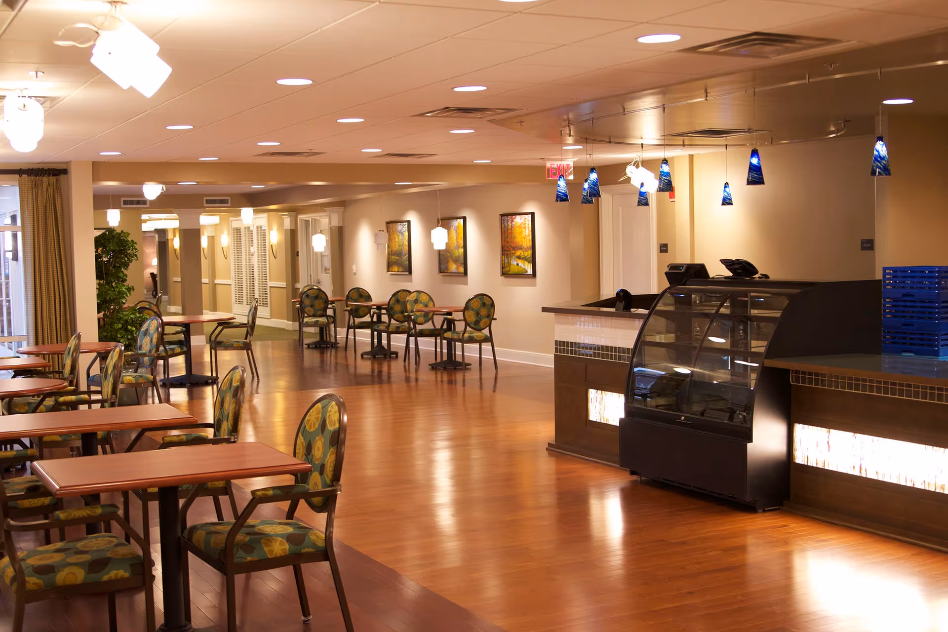 A spacious dining area in a senior living facility with wooden floors, multiple tables and chairs with patterned upholstery, a counter with a display case, and modern pendant lighting. The walls are decorated with framed artwork and there are plants and curtains near the windows.