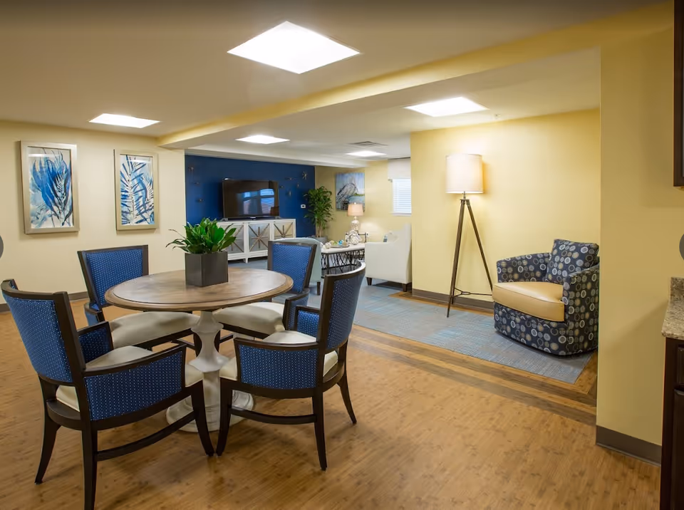 Bright common room with a round wooden table surrounded by blue chairs, a TV console, and a cozy seating area.