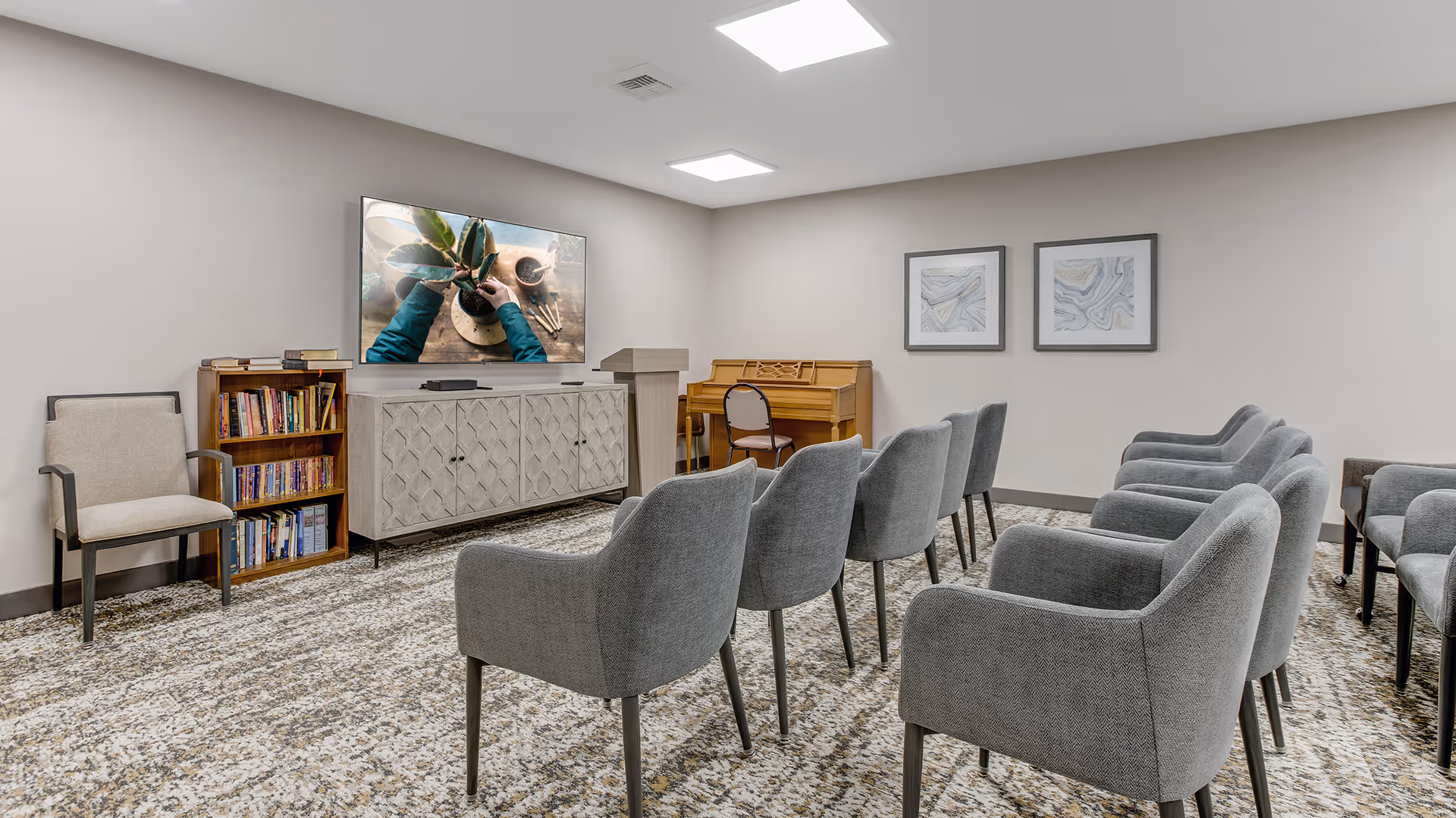 A small meeting or activity room with two rows of gray upholstered chairs facing a TV mounted on the wall. Below the TV is a cabinet and a small bookshelf filled with books. A wooden piano with a chair is positioned against the back wall, which also has two framed abstract artworks. The room has neutral-colored walls and carpeted flooring.