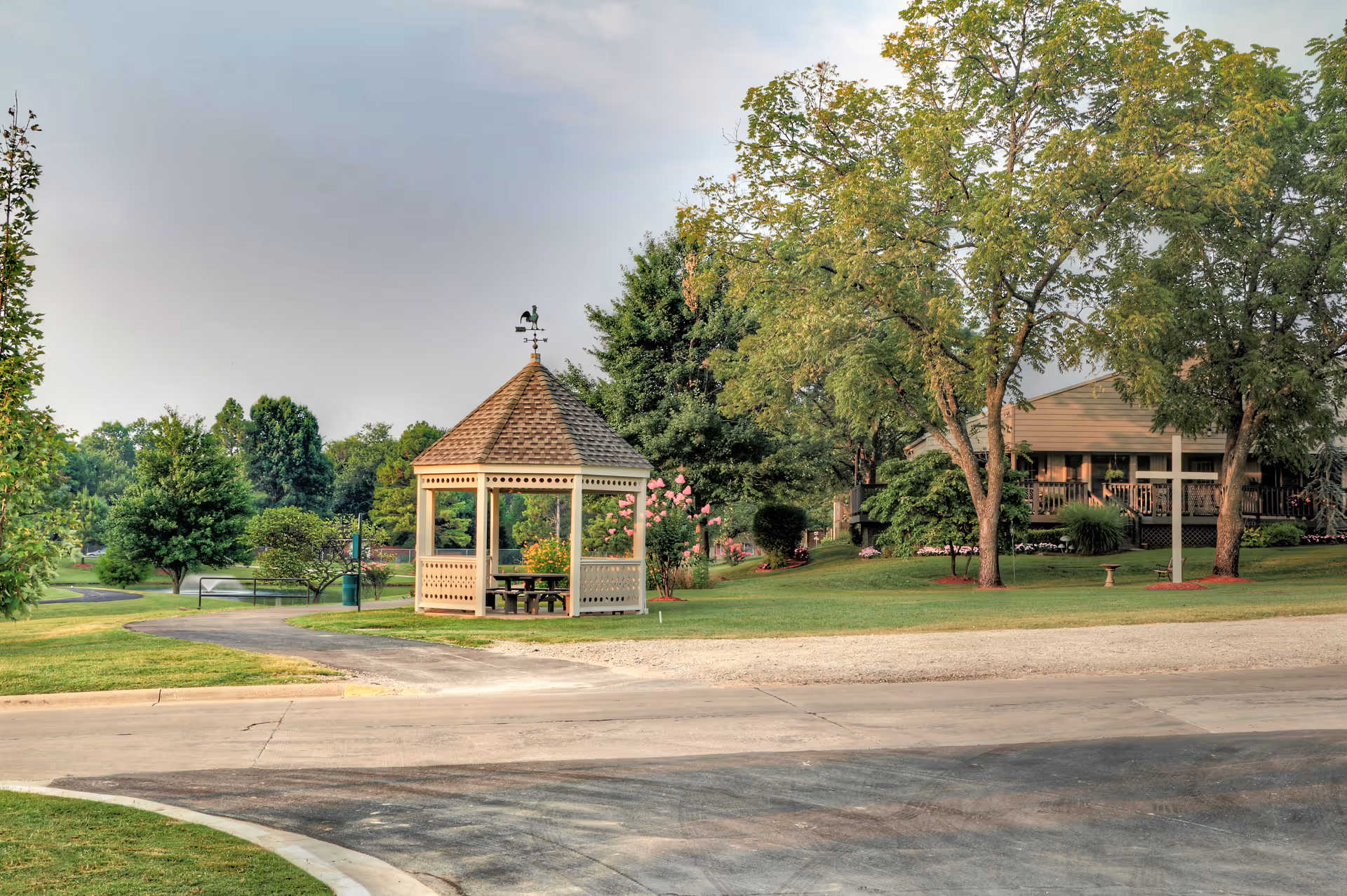 A peaceful outdoor scene at Trinity Woods featuring a small gazebo with a weather vane on top, surrounded by green grass, trees, and flowering bushes. A paved pathway curves around the gazebo, and a building with a porch is visible in the background.