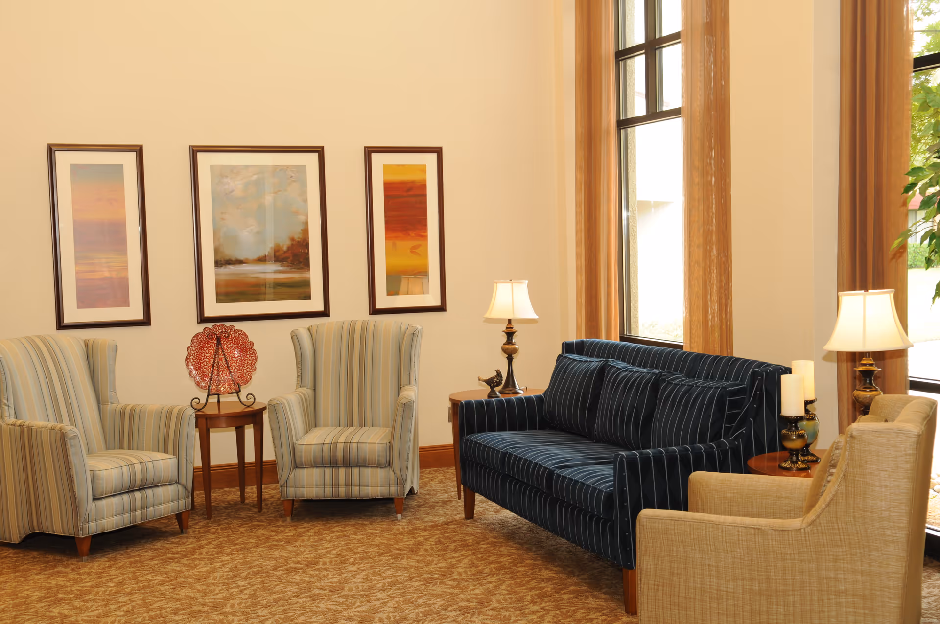 Sunlit seating area with upholstered chairs, a striped sofa, side tables, lamps, and framed artwork on the wall.