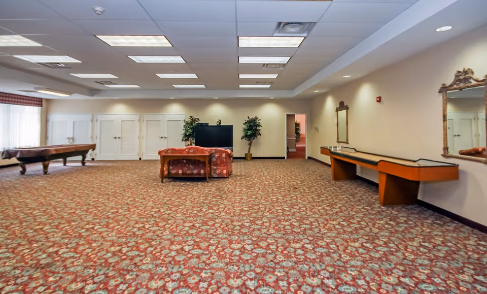 A large common room with patterned carpet, a pool table on the left, a floral couch facing a TV in the center, two potted plants behind the TV, and a shuffleboard table along the right wall with two ornate mirrors above it. The room has a drop ceiling with fluorescent lights and beige walls.