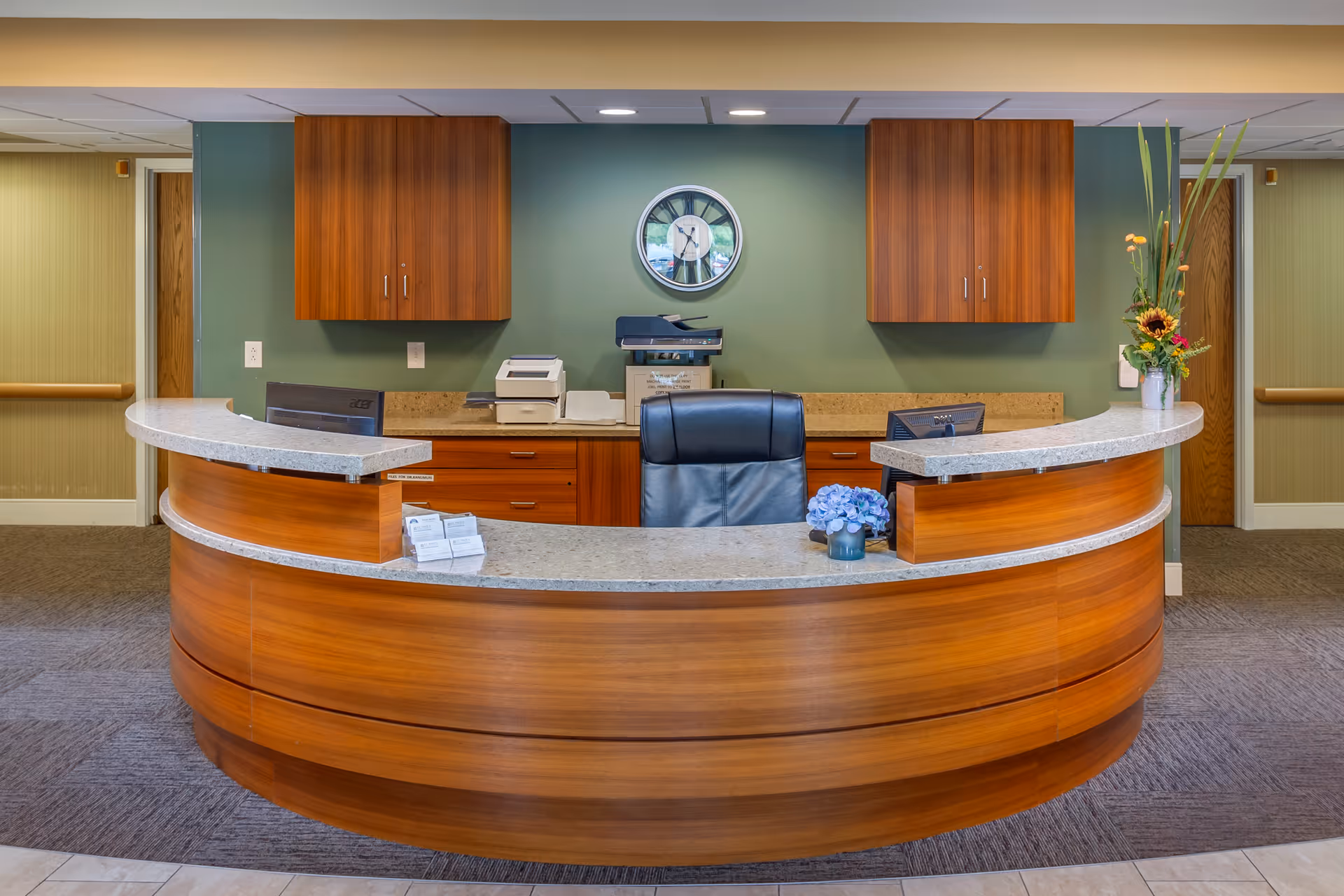 Curved wooden reception desk in a lobby with an office chair, cabinets, wall clock and a vase of flowers.
