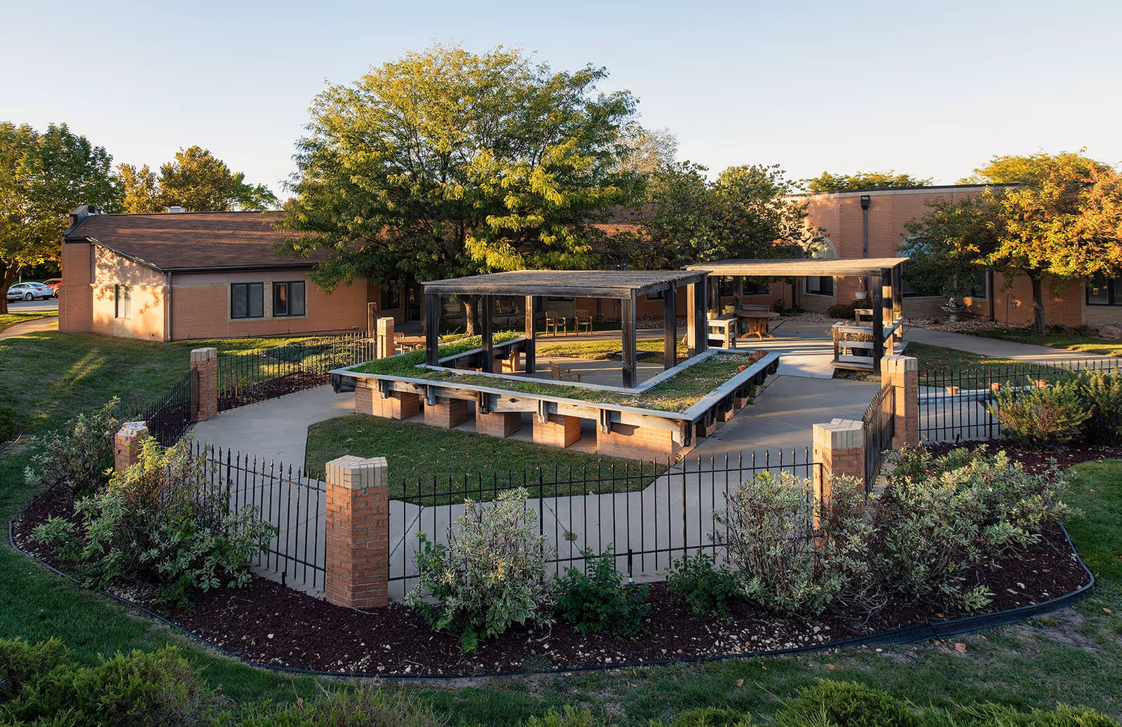 Outdoor courtyard area at Topeka Presbyterian Manor featuring a fenced garden space with brick pillars and black metal fencing, a concrete walkway, two shaded pergola structures with seating underneath, surrounded by green grass, shrubs, and trees with a building in the background.