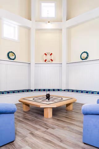 A bright, nautical-themed seating area with a hexagonal wooden table featuring a compass design in the center. The room has white paneled walls with built-in bench seating along the walls, topped with blue and green plaid cushions. Two blue armchairs are partially visible in the foreground. The walls have small windows near the ceiling and decorative life rings mounted on the walls.
