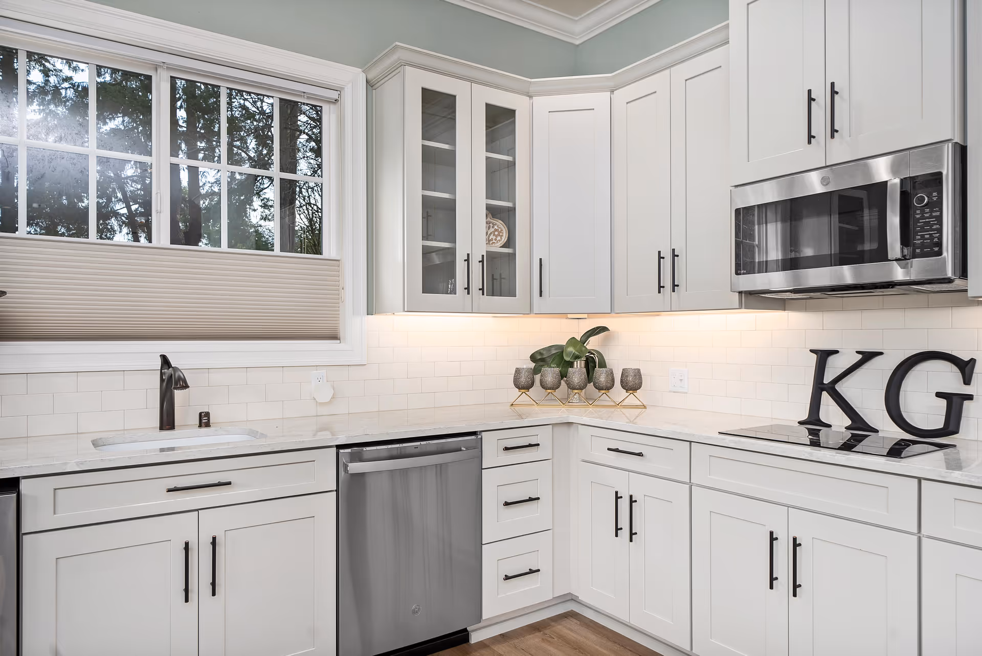 Bright modern kitchen with white cabinets, stainless steel appliances, a sink under a window, and decorative 'KG' letters on the counter.
