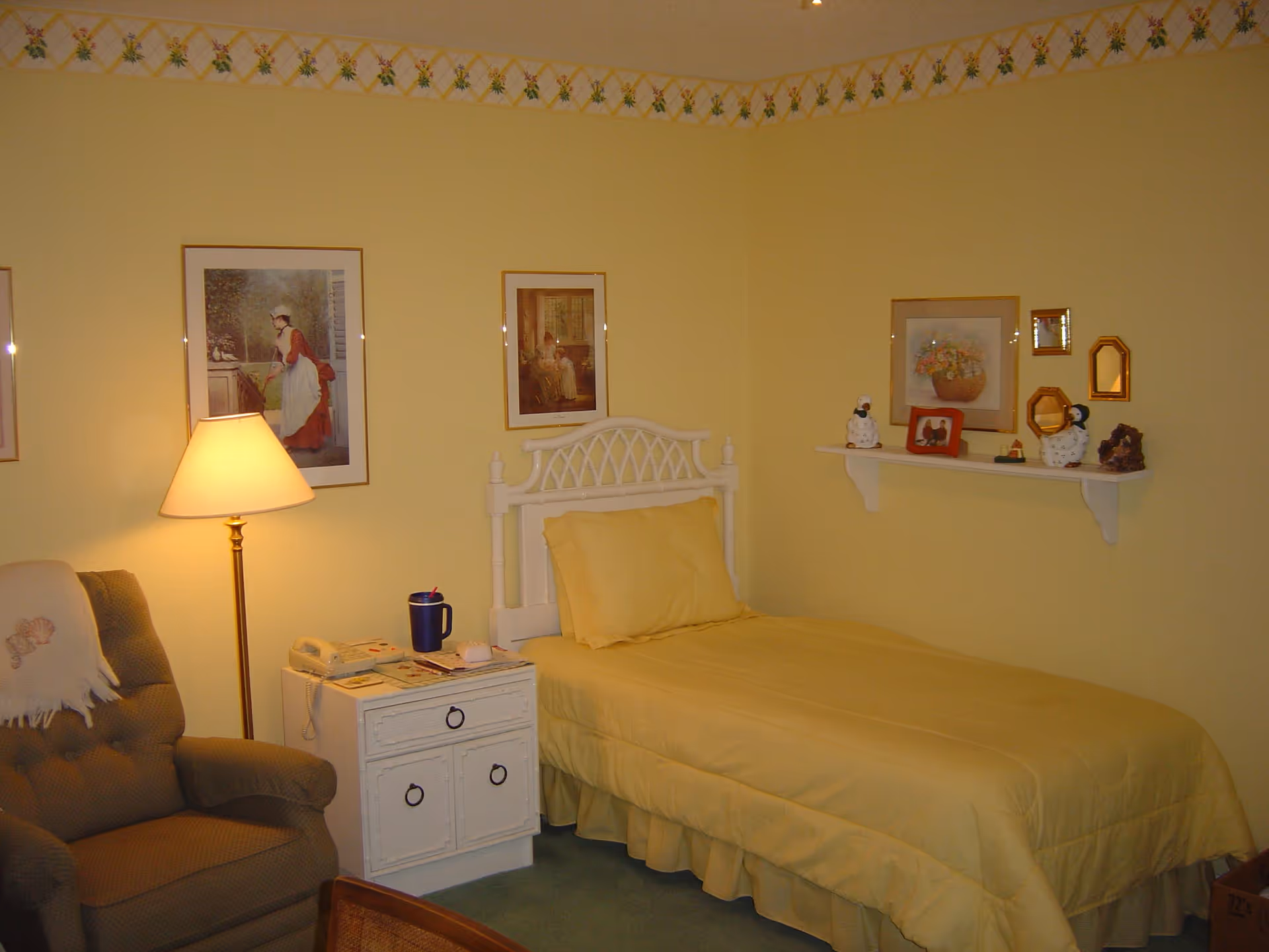 A small yellow-themed bedroom with a single bed, nightstand, lamp, armchair and framed pictures on the walls.