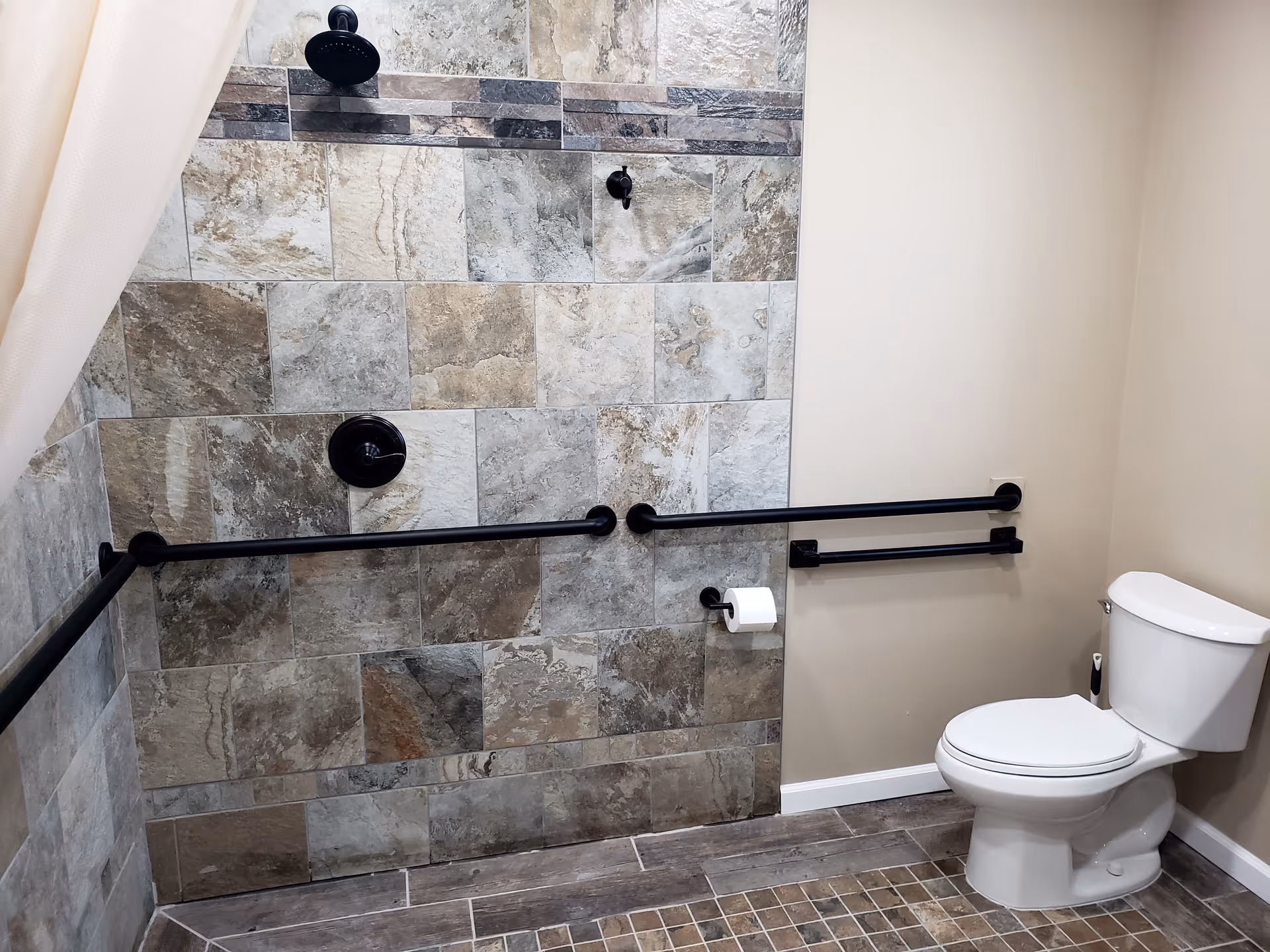 A bathroom with a tiled walk-in shower featuring a black showerhead and grab bars. There is a white toilet with a toilet paper holder mounted on the wall next to it. The walls are painted beige and the floor has brown and beige tiles.